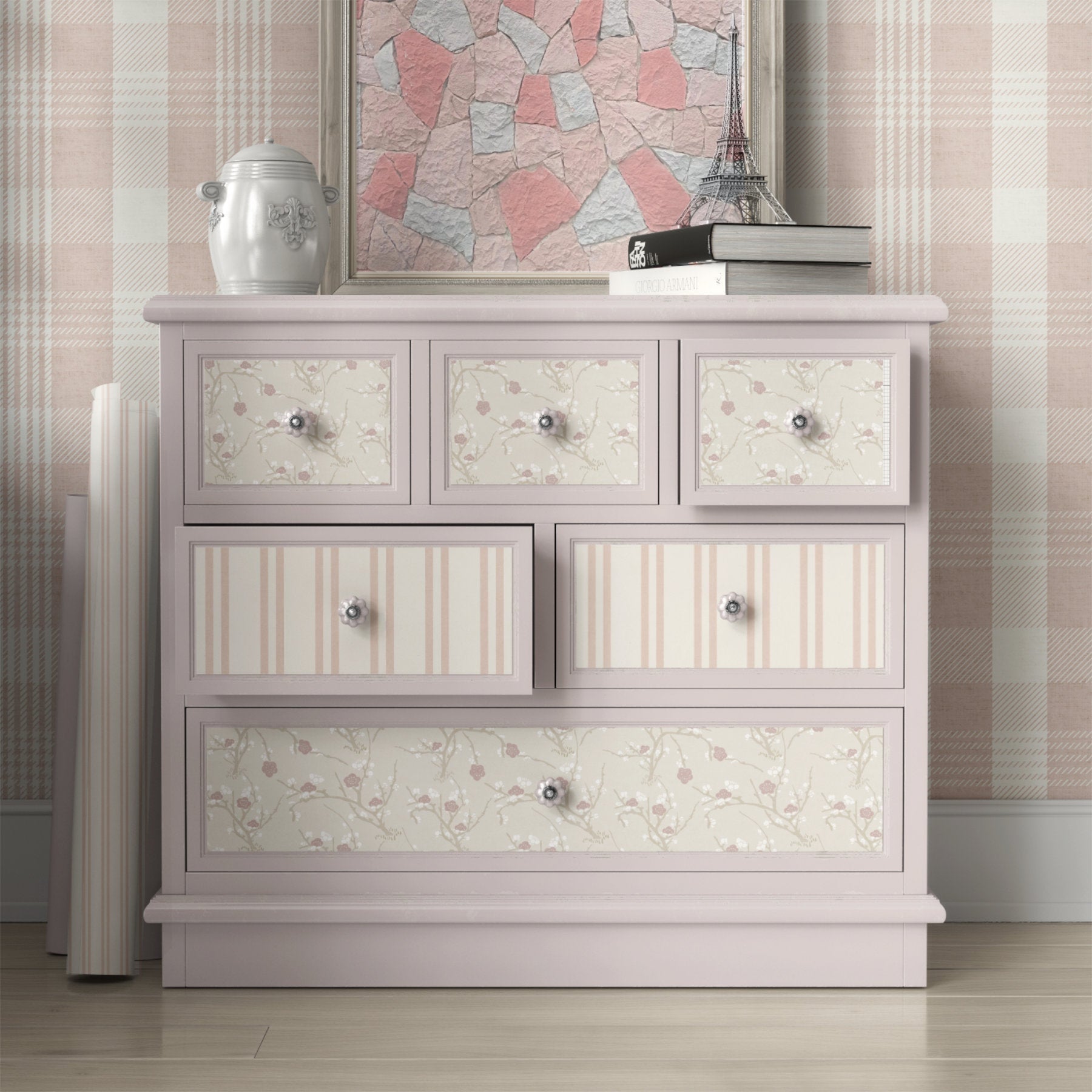 A light pink wooden dresser with six drawers, featuring floral and striped wallpaper patterns on the drawer fronts, styled in a bedroom setting with wallpaper and decorative items on top.