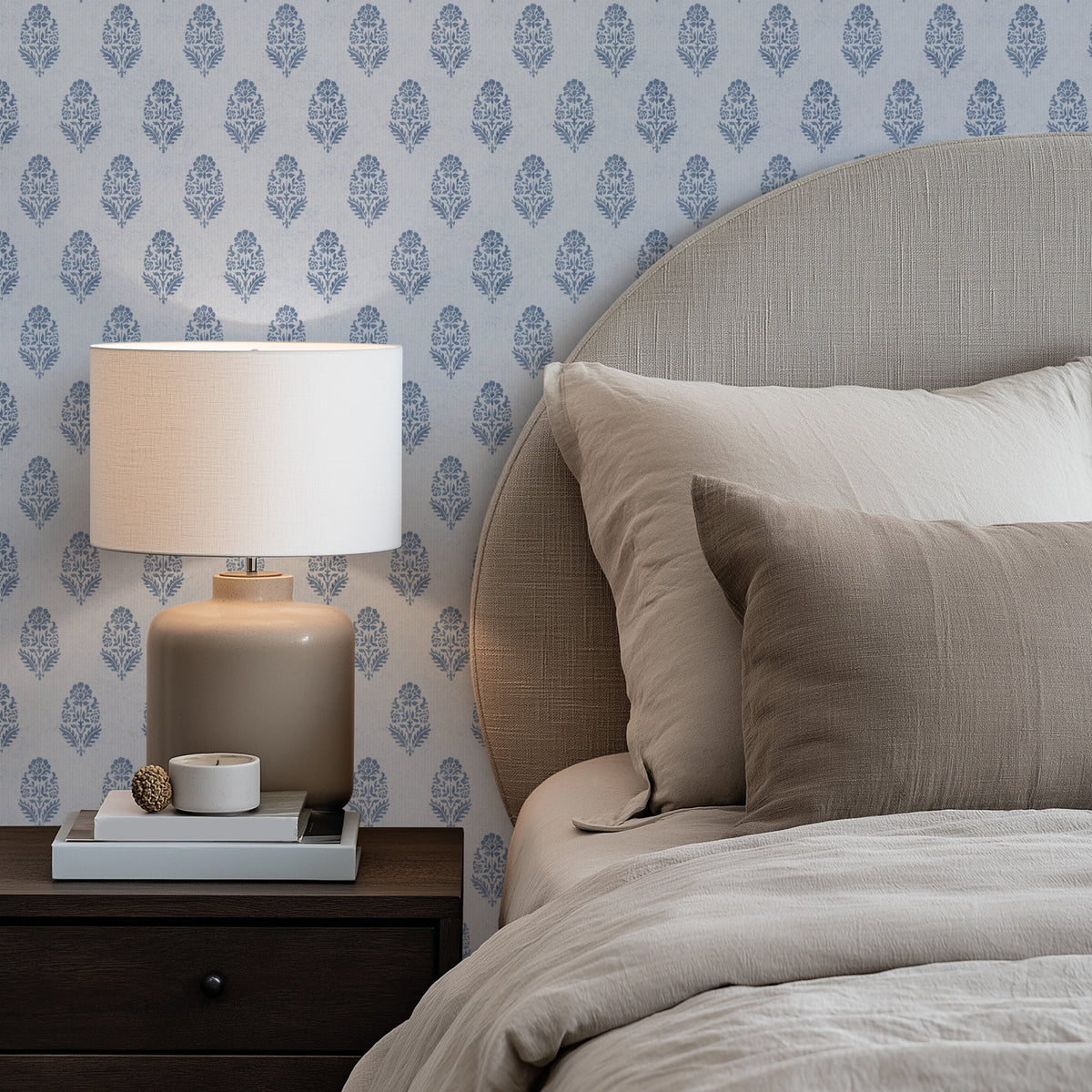 A styled bedroom corner featuring a bed with neutral beige bedding and pillows, a matching upholstered headboard, a lit table lamp on a dark wood nightstand, and blue and white floral block print wallpaper in the background.