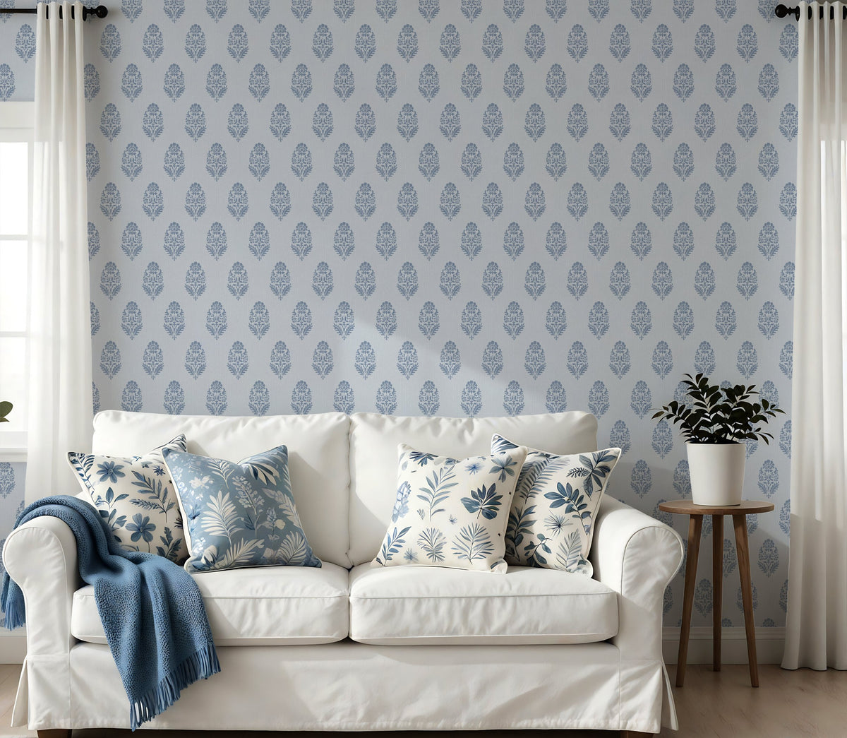 A coastal-style living room with a white sofa, blue and white cushions and a blue throw over the left arm. A small plant sits on a side table on the right, and the wall behind is covered in blue floral block print wallpaper.