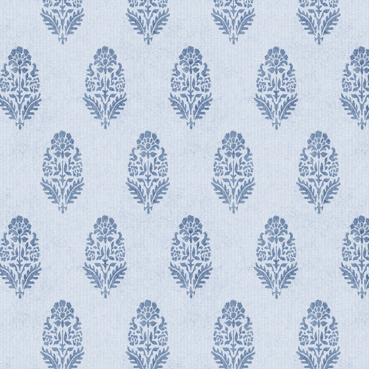 A close-up of denim blue floral block print wallpaper on a pale blue-white background. Small stylised bouquets of petals and leaves repeat in softly offset rows, with a slightly inky, textured edge.