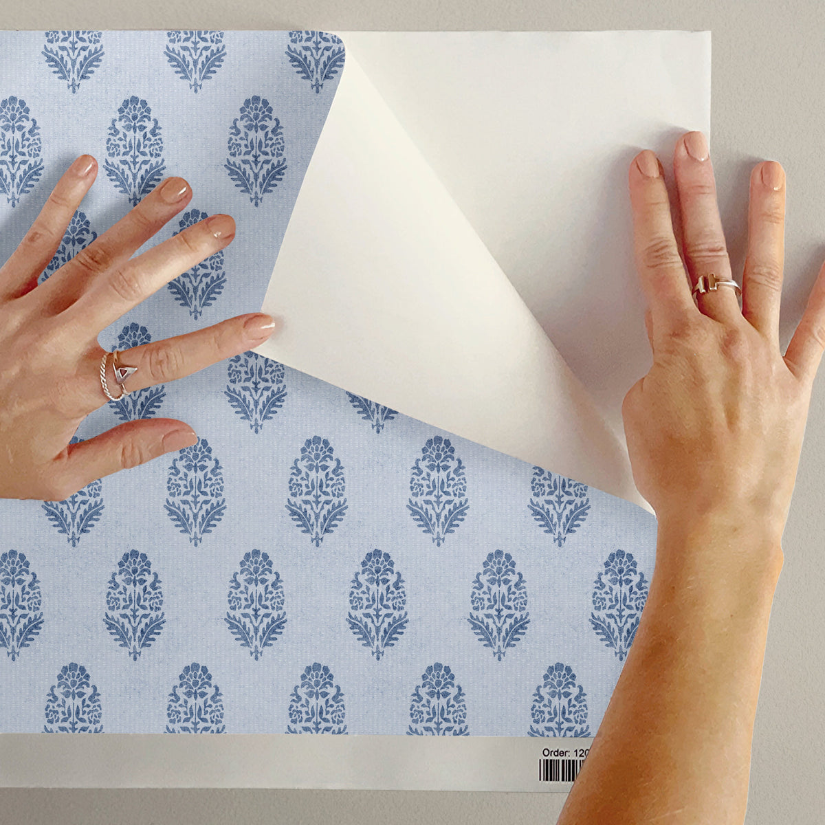 Two hands peel the white backing paper from a peel and stick sample of blue floral block print wallpaper held against a pale wall. The small stylised flower pattern shows on the left, with the backing curling up on the right.