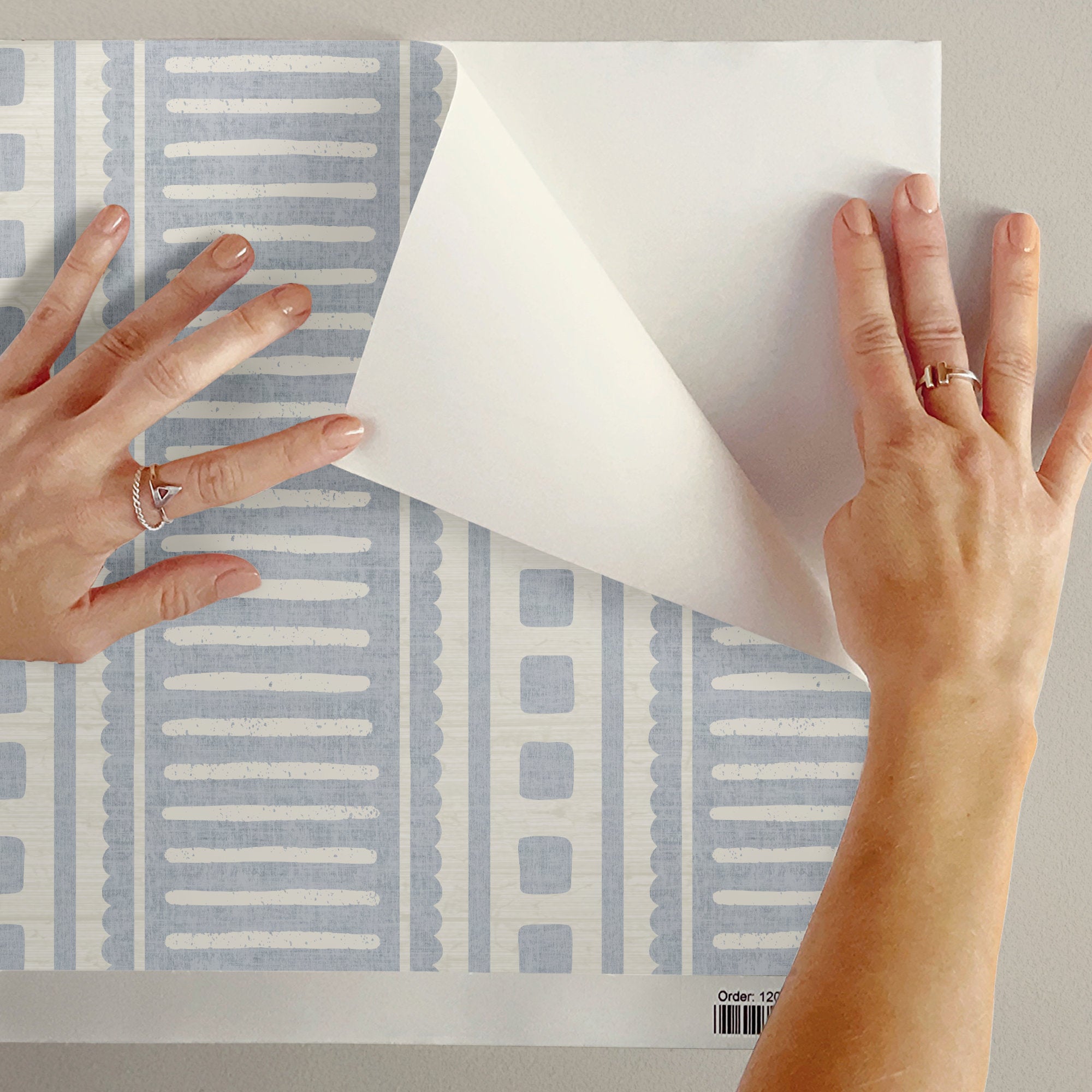 Hands peeling back the backing of cornflower blue peel and stick coastal stripe wallpaper