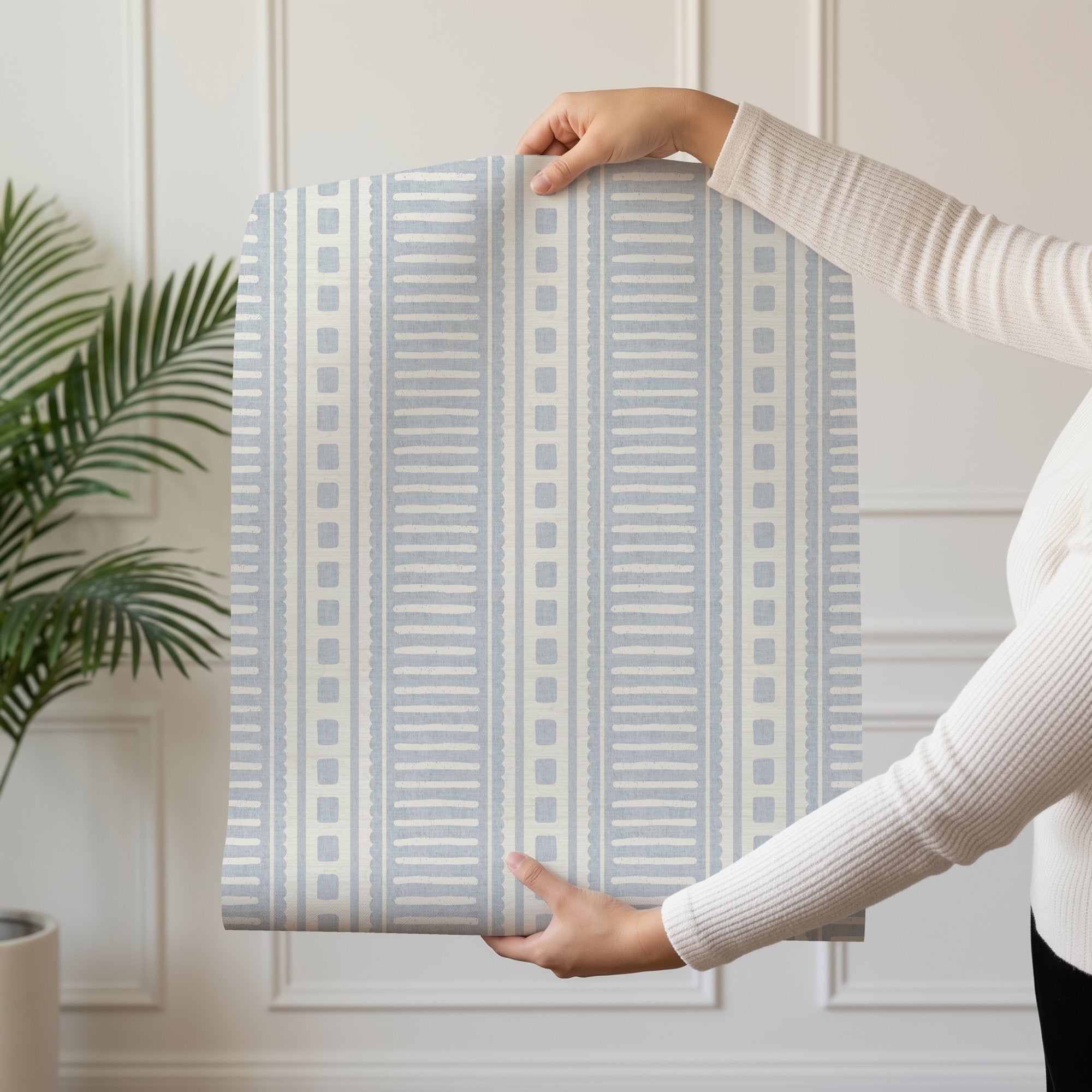A person holding up a roll of cornflower blue coastal stripe wallpaper, showing the block-print stripe pattern