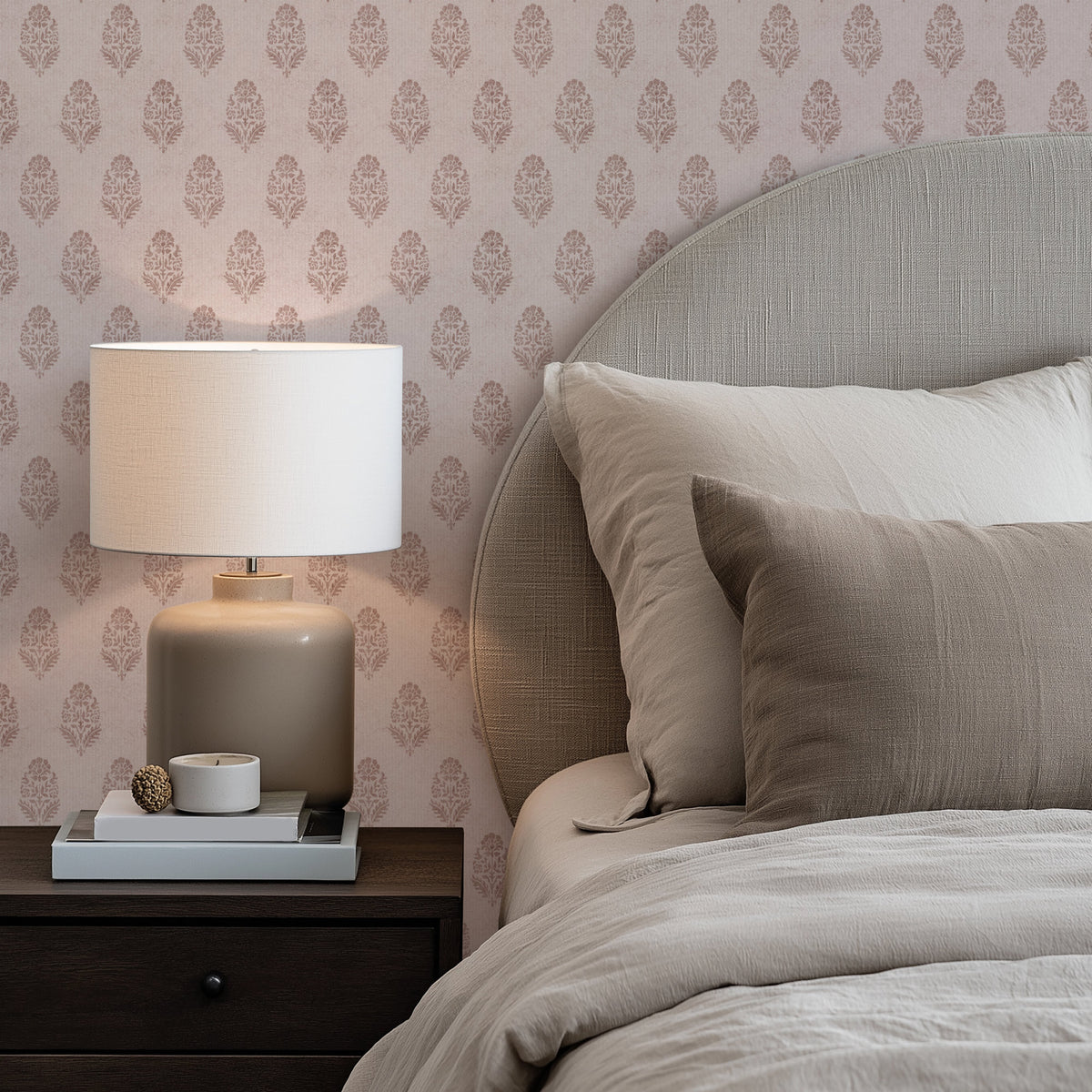A bedroom corner with a bed in neutral beige bedding and a round-topped upholstered headboard, set against dusty pink floral block print wallpaper. On the left, a table lamp with a white shade stands on a dark wood nightstand beside a small stack of books.