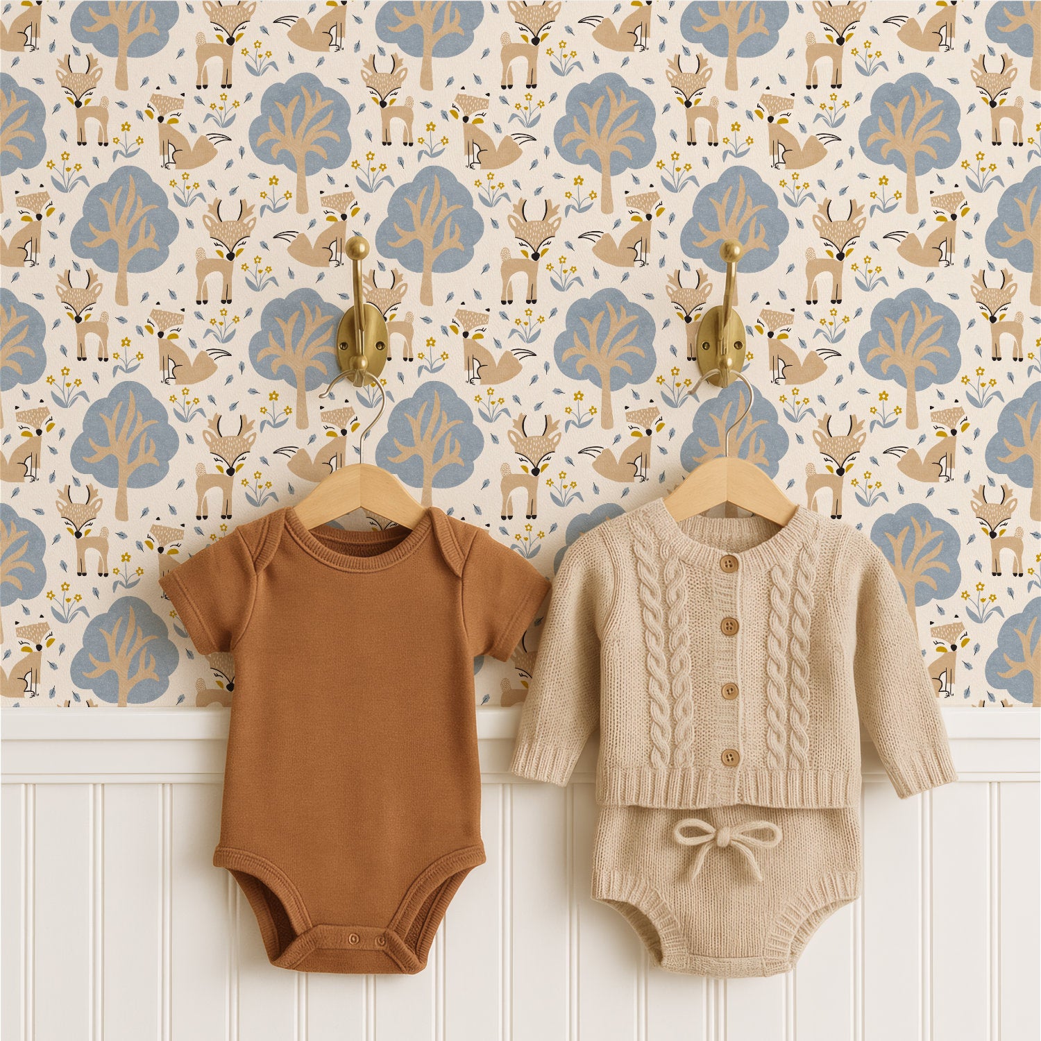 Two baby onesies hanging on wooden hangers against a woodland nursery wallpaper pattern . The left oneie is a solid warm camel brown, and the right oneie is a cream-colored cable-knit cardigan with matching knit shorts.
