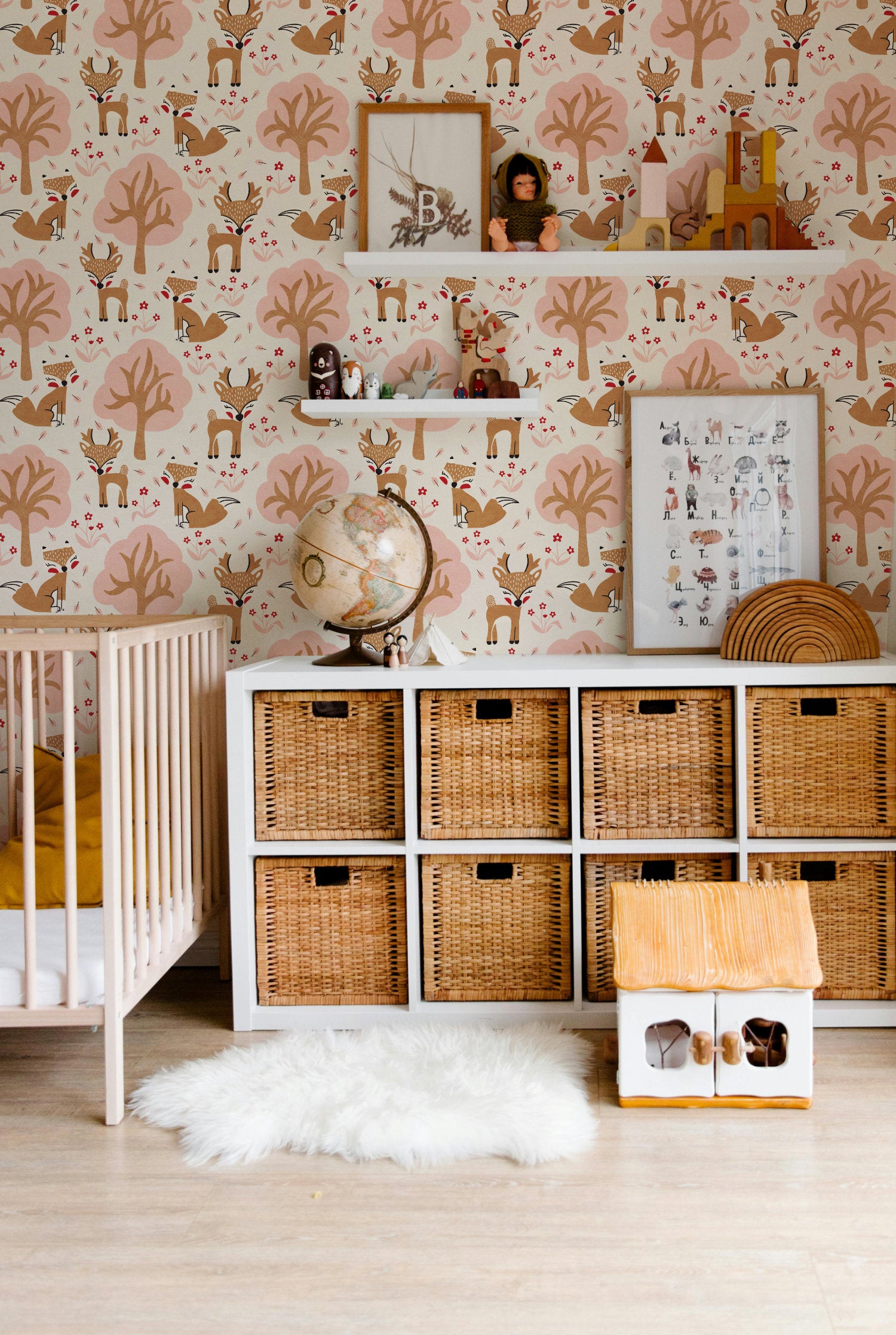 A styled nursery interior featuring a crib, white storage cabinet with woven wicker bins, a white fluffy rug, and a small wooden house. The wall is covered in pink and beige woodland nursery wallpaper pattern featuring foxes and deer