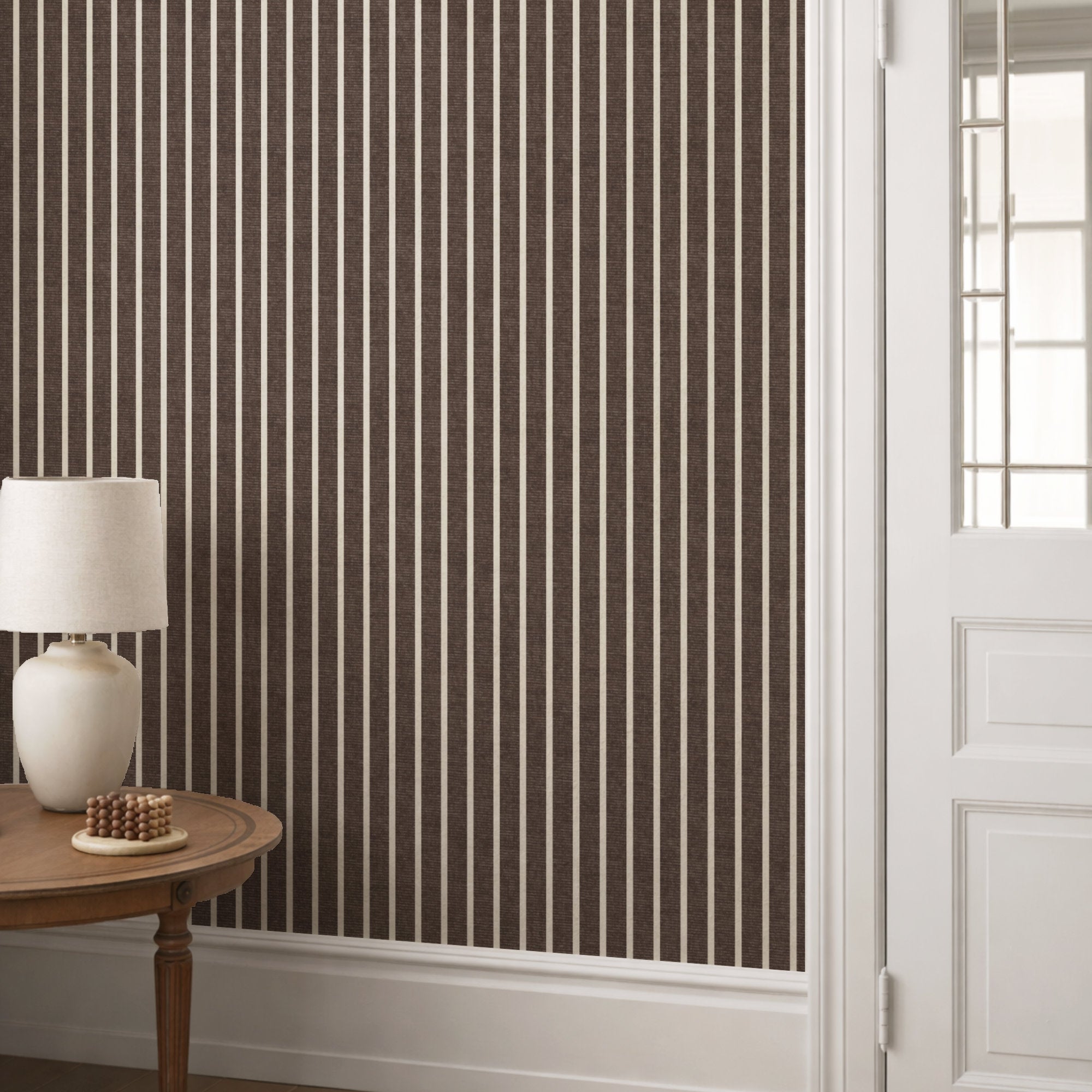 Dark brown pinstripe wallpaper in a hallway behind a round console table with a cream lamp