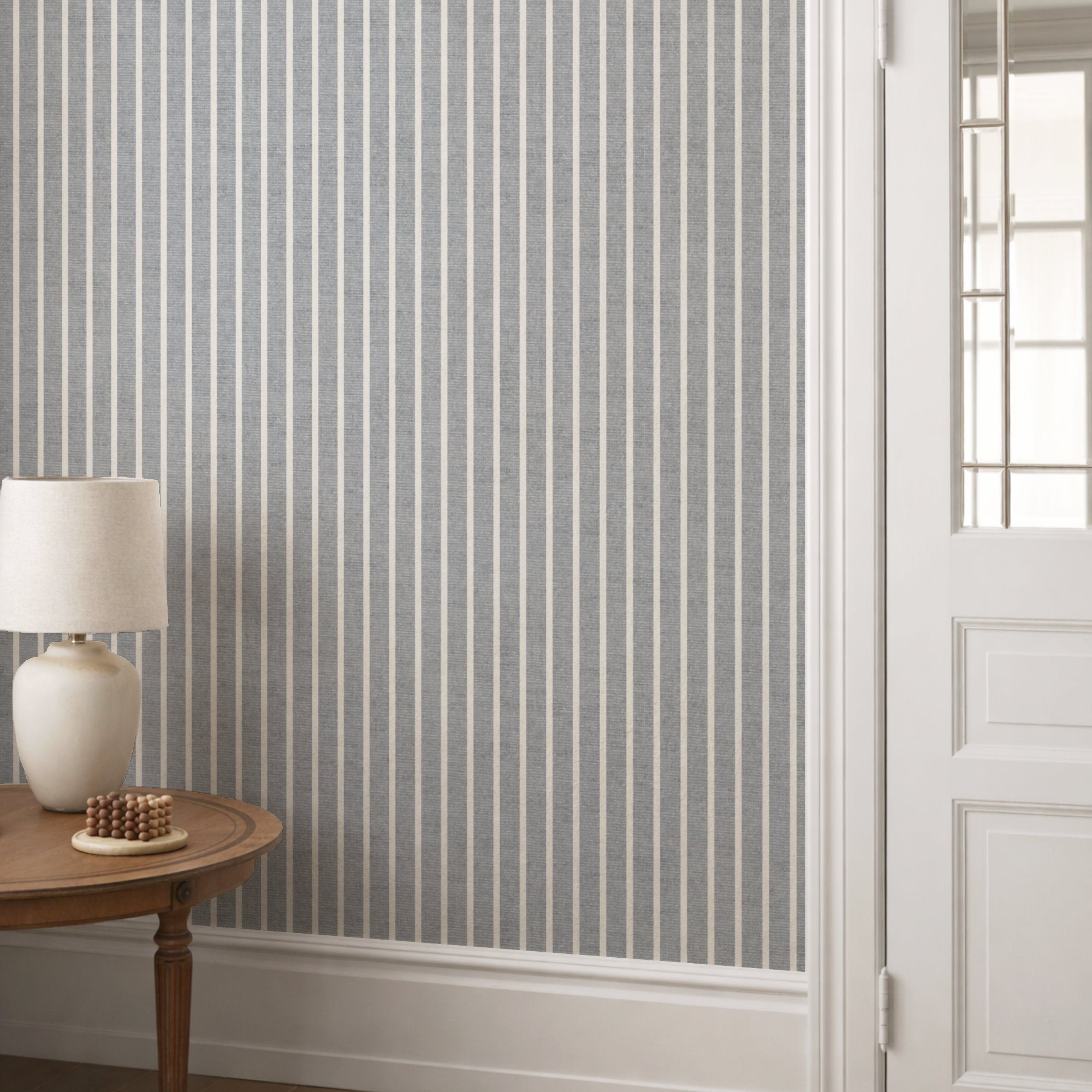 Light grey pinstripe wallpaper in a hallway behind a round console table with a cream lamp and panelled door