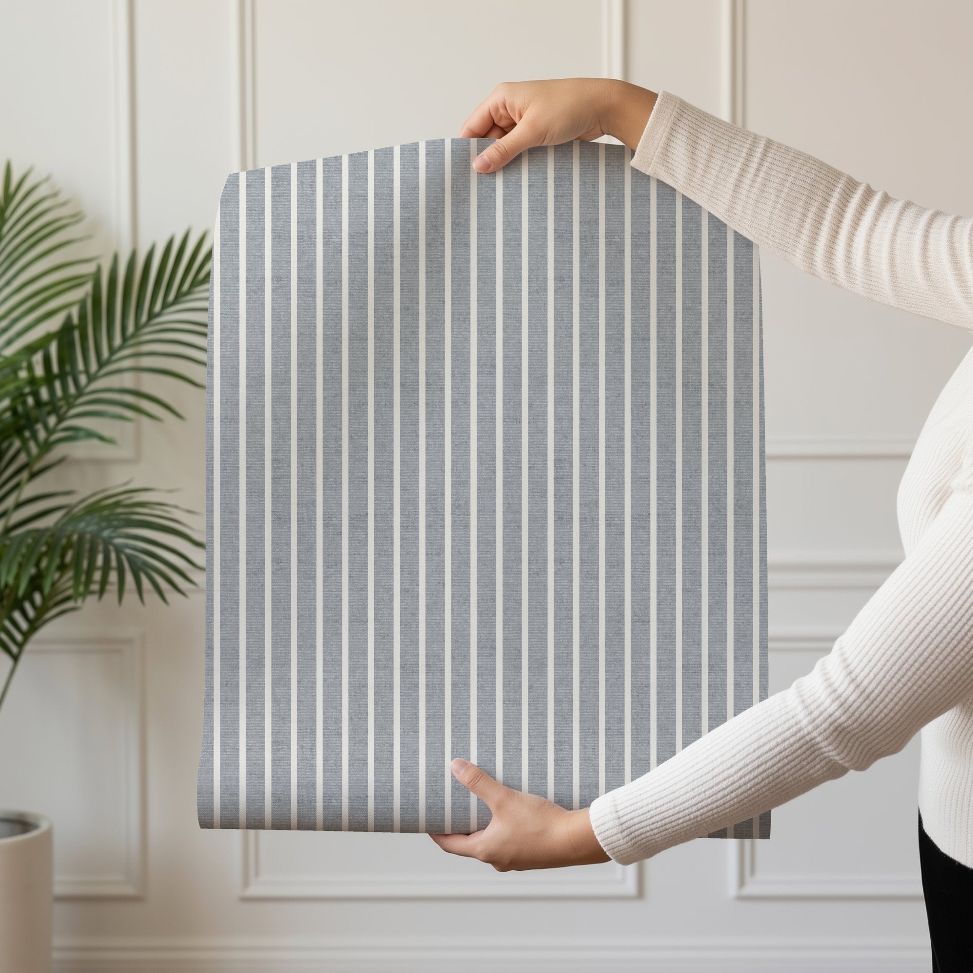 A person holding up a roll of light grey pinstripe wallpaper beside a palm plant