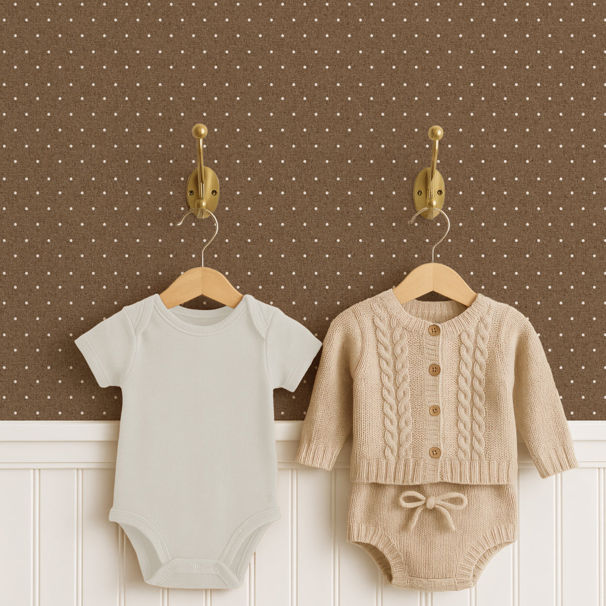 Two brass hooks hold a pale cream baby bodysuit and an oatmeal cable-knit cardigan with matching knitted shorts on wooden hangers, above white panelled wainscoting. The wall behind is espresso brown polka dot wallpaper scattered with small cream dots.