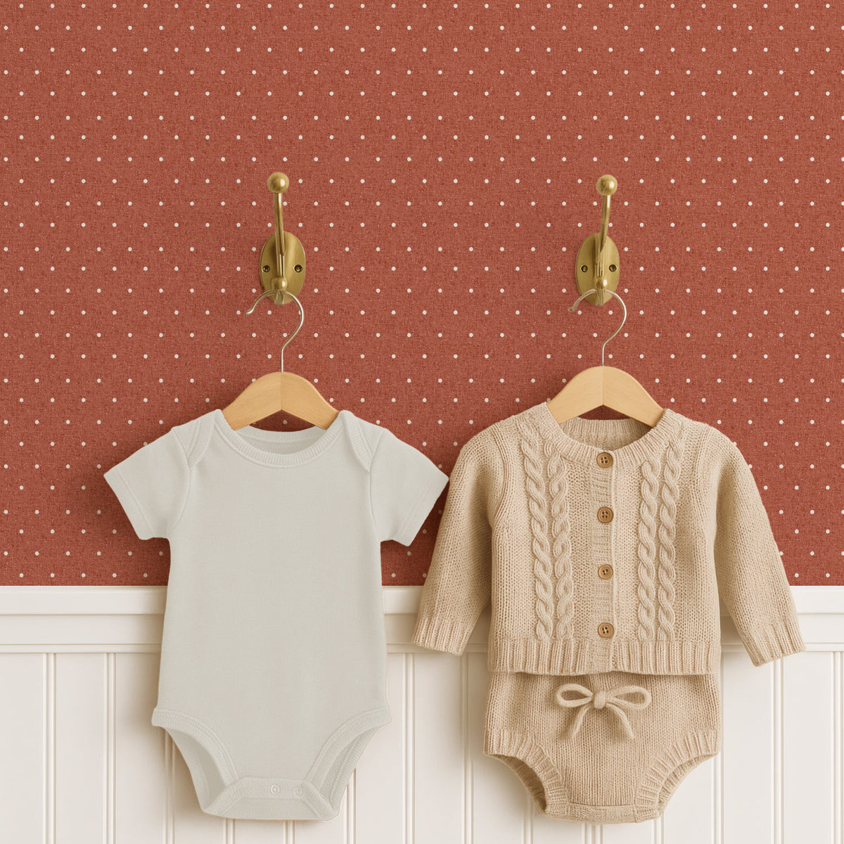 A pale cream baby bodysuit and an oatmeal cable-knit cardigan with matching knitted shorts hang on wooden hangers from two brass hooks, above white panelled wainscoting. Behind them, terracotta polka dot wallpaper carries small cream dots on a linen-look ground.