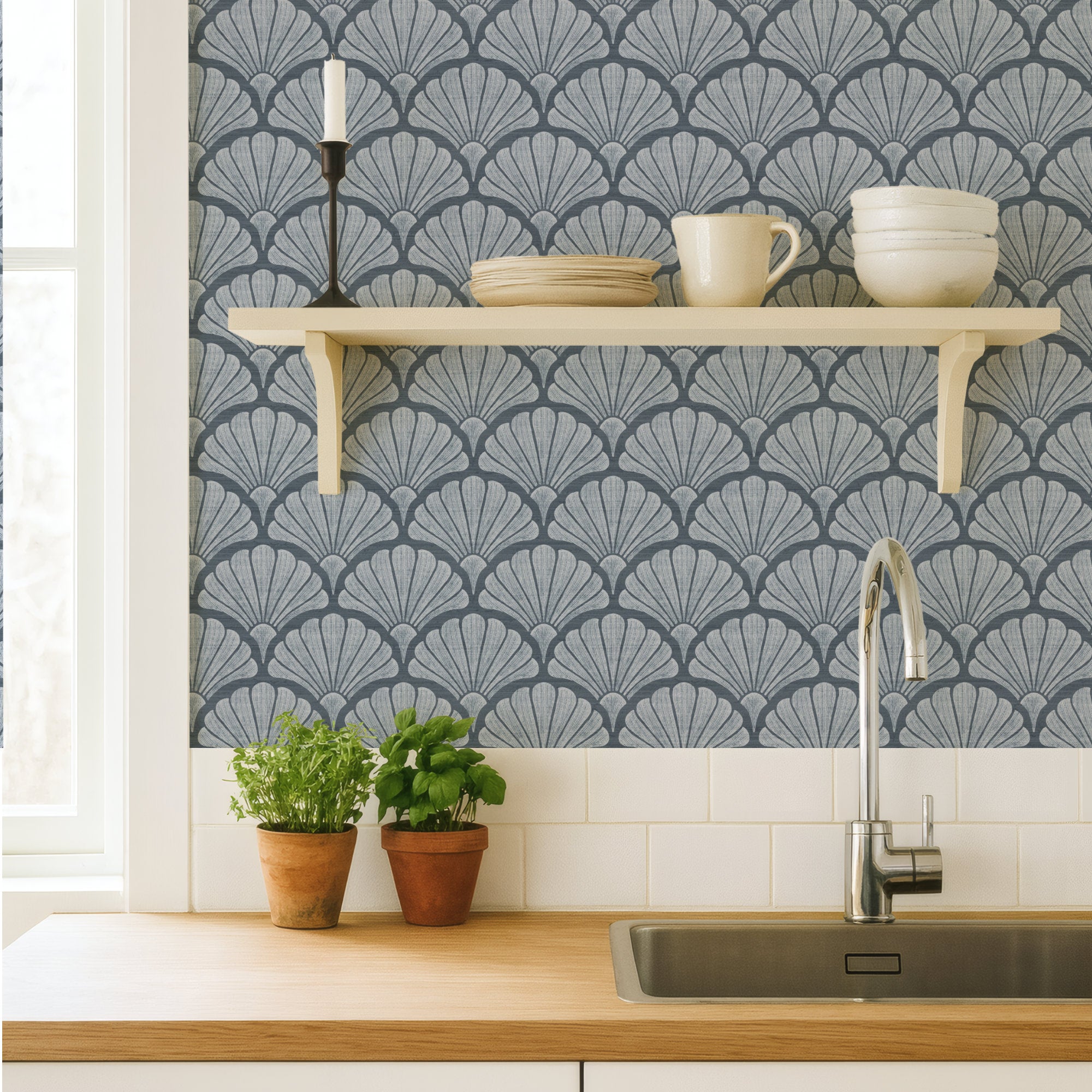 Denim blue scallop shell wallpaper in a kitchen, bold hand-drawn fan shells behind a cream shelf of crockery, above a wood worktop, sink and potted herbs