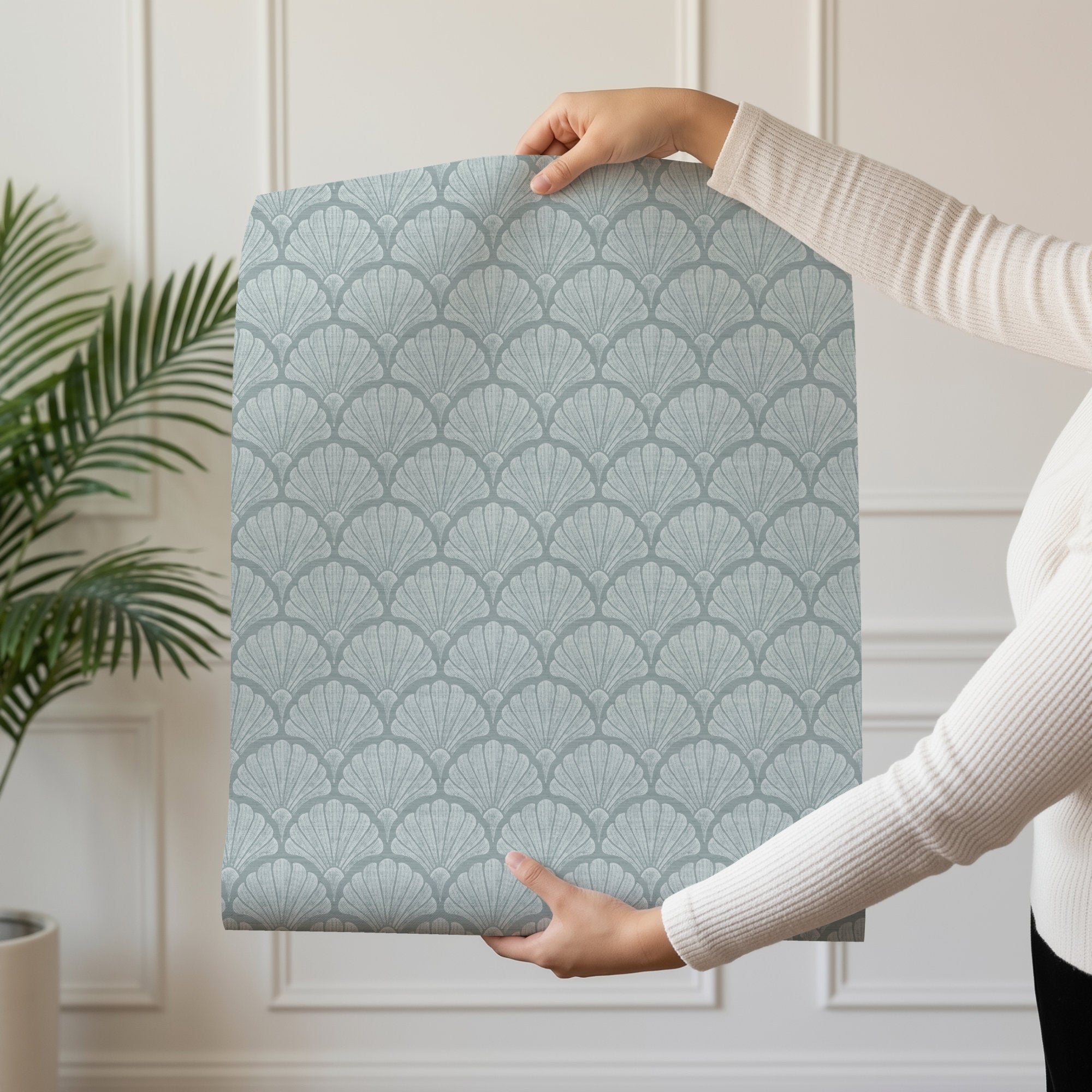 A person holding up a panel of light blue scallop shell wallpaper by a window, showing the hand-drawn fan shell pattern