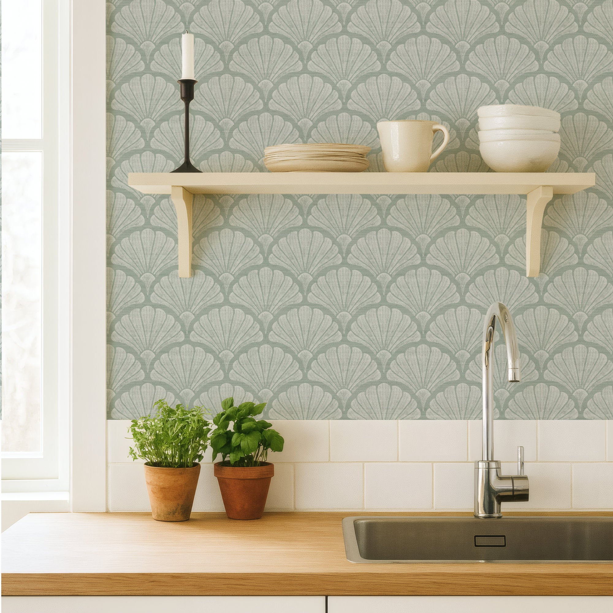 Sage green scallop shell wallpaper in a kitchen, hand-drawn fan shells behind a cream shelf of crockery, above a wood worktop, sink and potted herbs