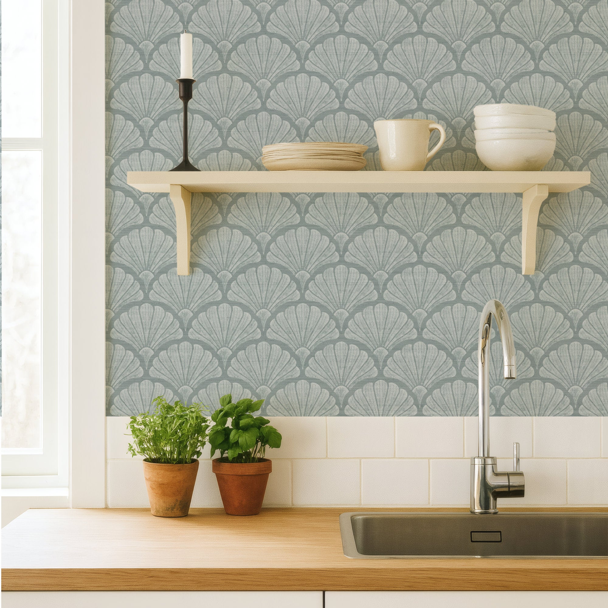 Light blue scallop shell wallpaper in a coastal kitchen, hand-drawn fan shells behind a cream shelf of crockery, above a wood worktop, sink and potted herbs
