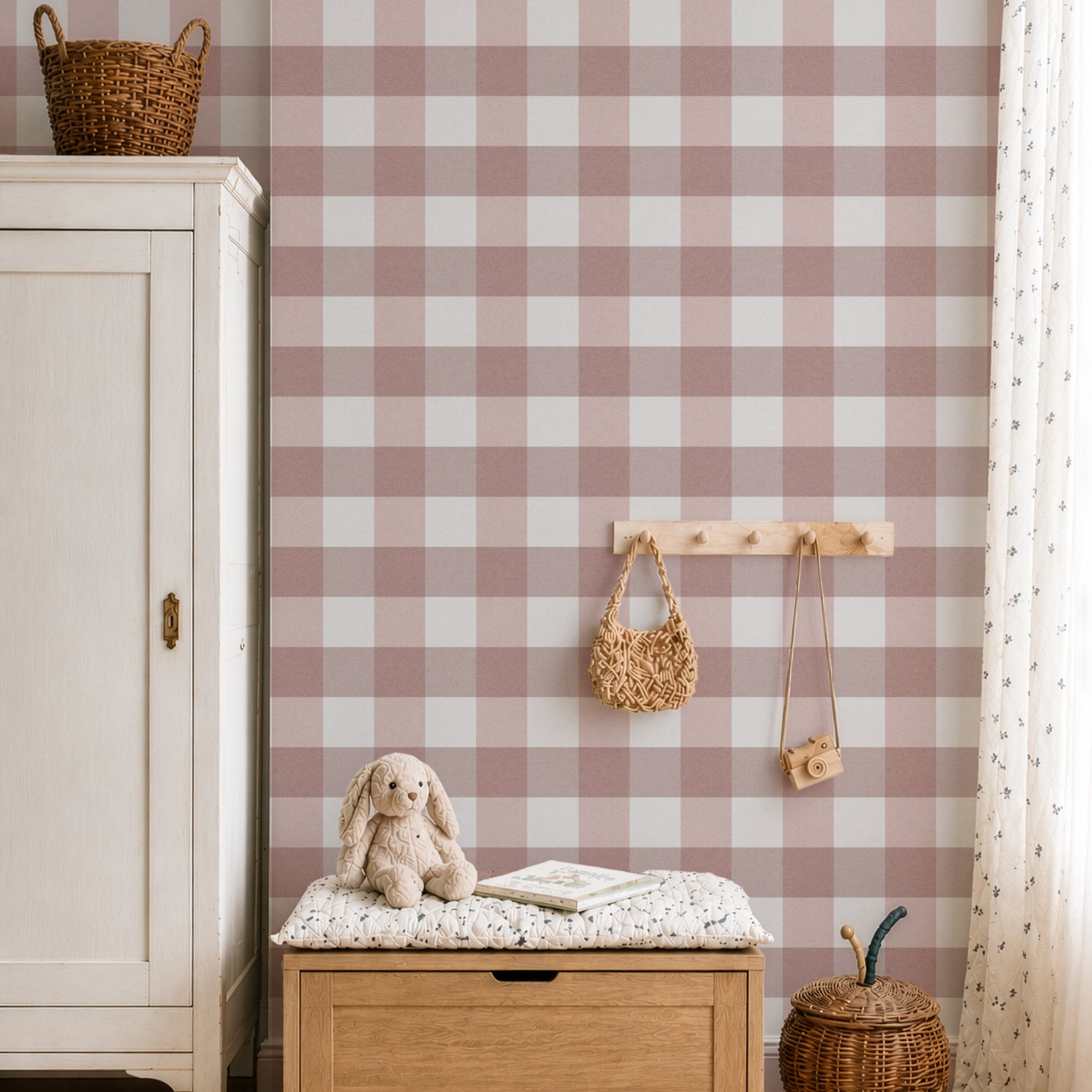 A styled nursery or playroom corner featuring dusty pink and white gingham wallpaper. In the foreground, a wooden storage bench with a floral cushion holds a plush bunny toy and a book, next to a light wood cabinet.