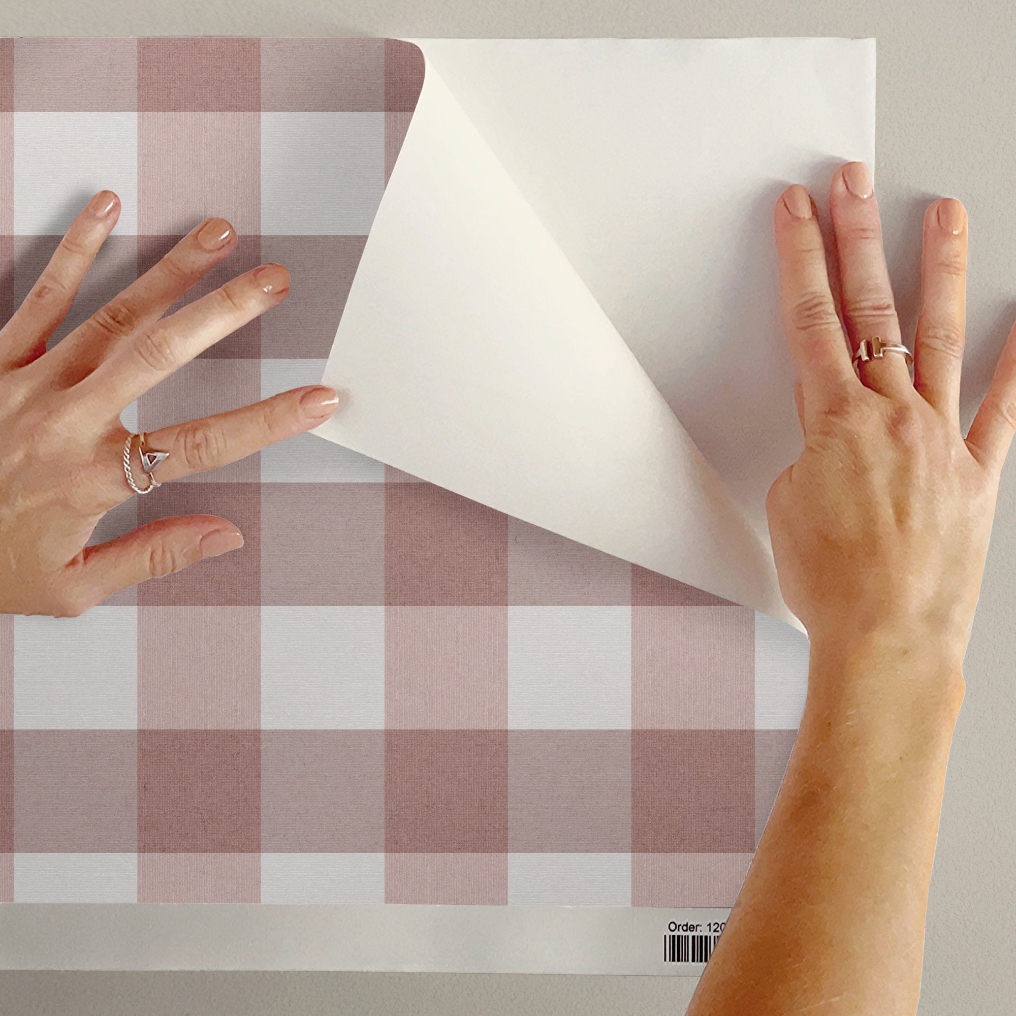 A person's hands peel back a corner of a sheet peel and stick wallpaper featuring a dusty pink and white gingham wallpaper pattern, showing the white backing.