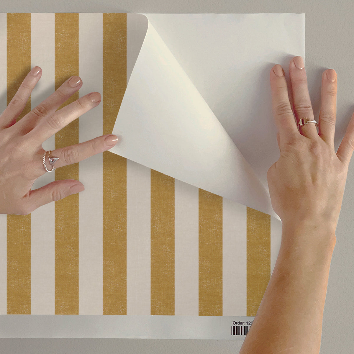 Hands peeling and applying peel and stick yellow wide stripe wallpaper in ochre yellow, easy to apply removable wallpaper
