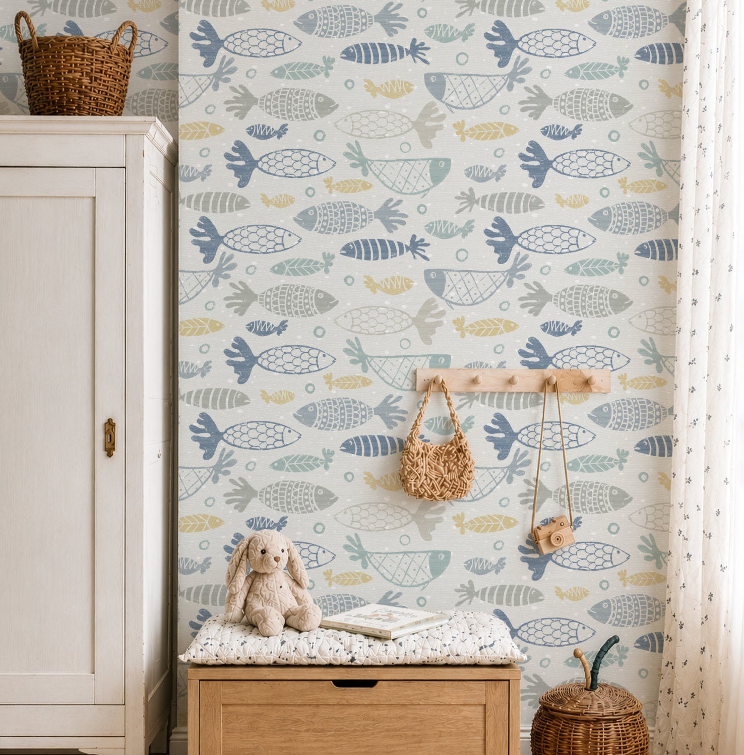 A styled nursery room corner in multicolour school of fish wallpaper, the hand-drawn fish in muted blue, grey and mustard on white. In front, a light wood toy chest, a cream fabric storage bin of plush toys, a small book shelf and a woven rug.