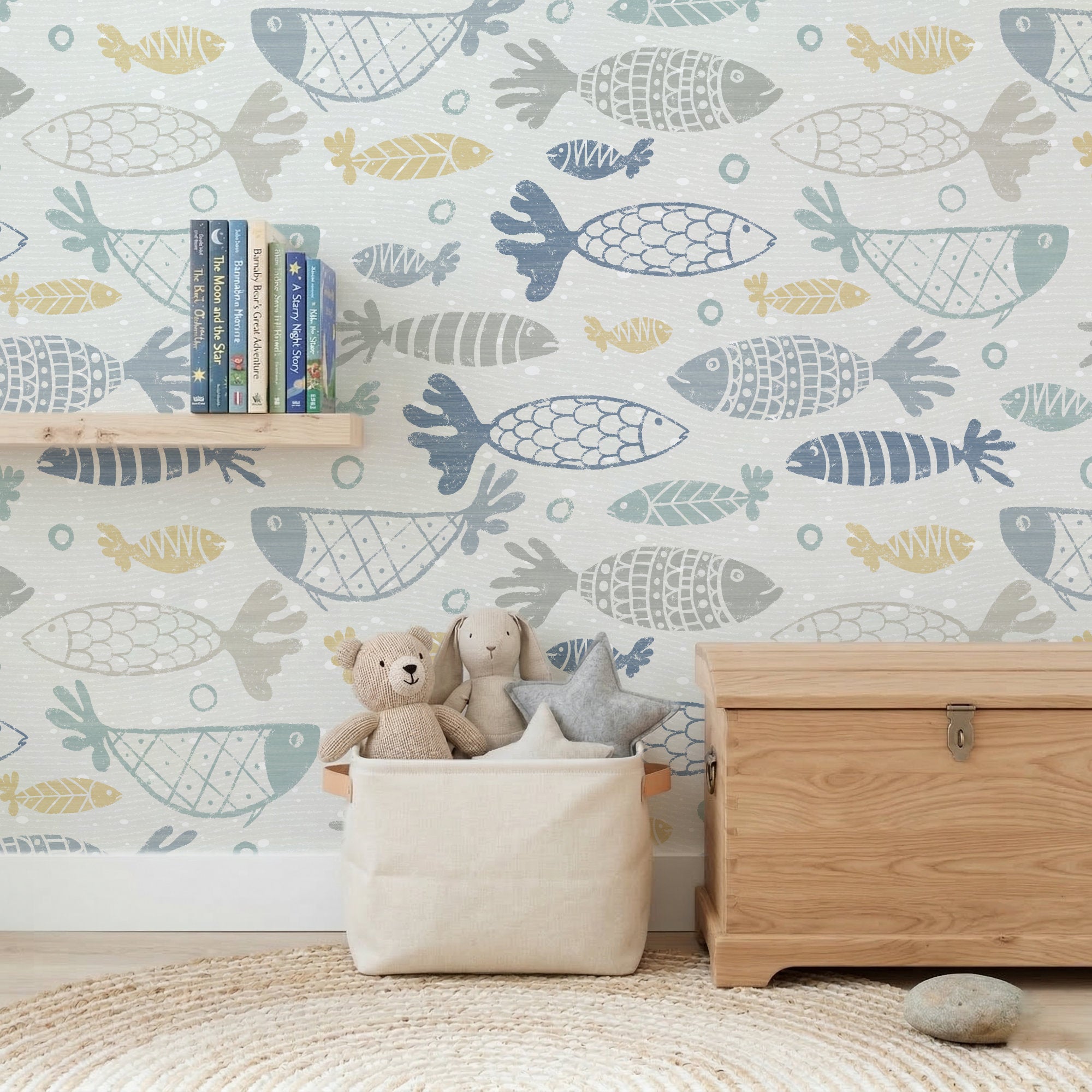 A children's room corner in multicolour school of fish wallpaper, the hand-drawn fish in muted blue, grey and mustard on white. In front, a light wood toy chest, a cream fabric storage bin of plush toys, a small book shelf and a woven rug.