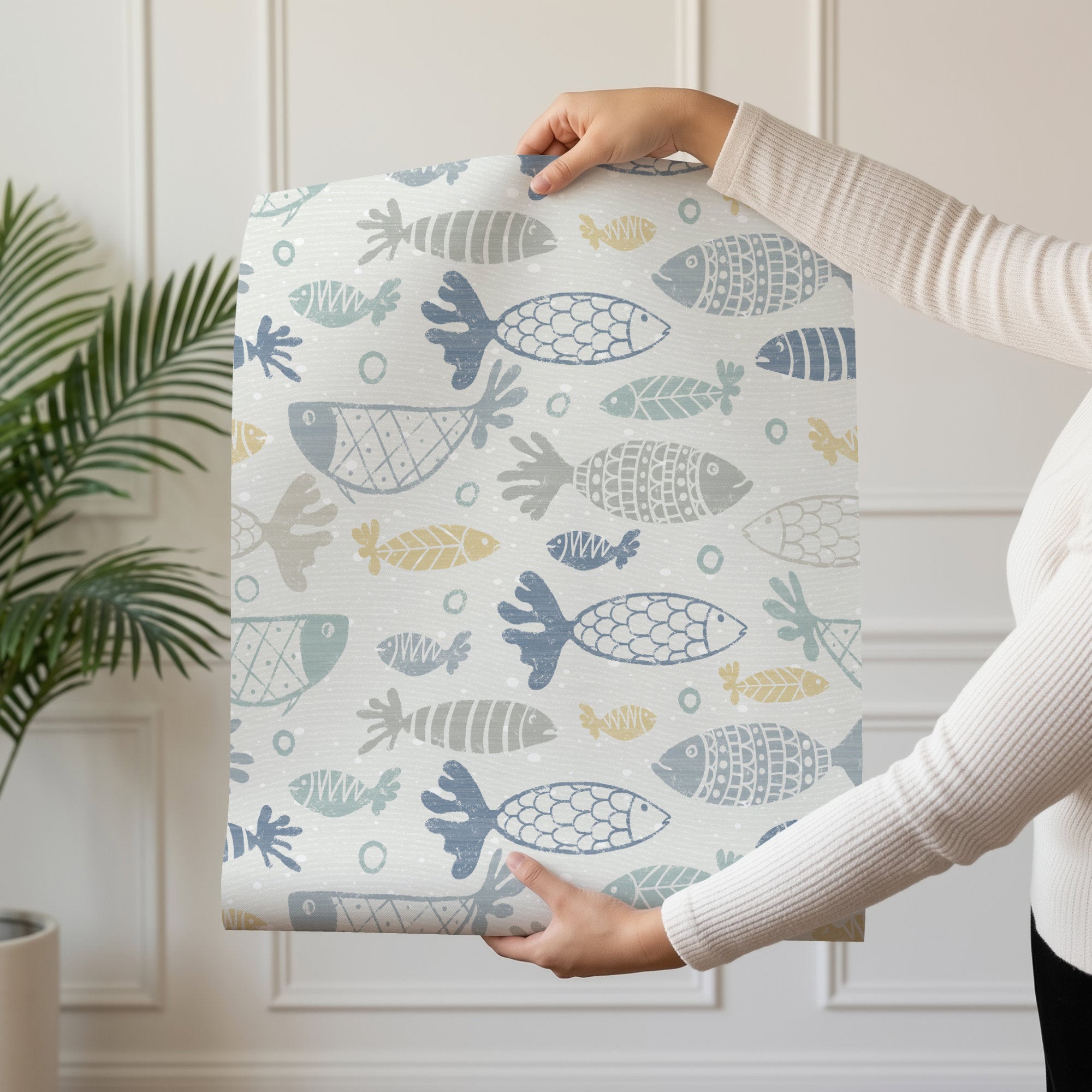A person holding up a panel of multicolour school of fish wallpaper, hand-drawn fish in blue, green, grey and mustard swimming in rows across a soft cream ground.