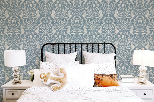 Blue seahorse wallpaper behind a white coastal bedroom setup with a black metal bedframe, textured bedding, and gold anchor cushion – calm coastal interior inspiration.