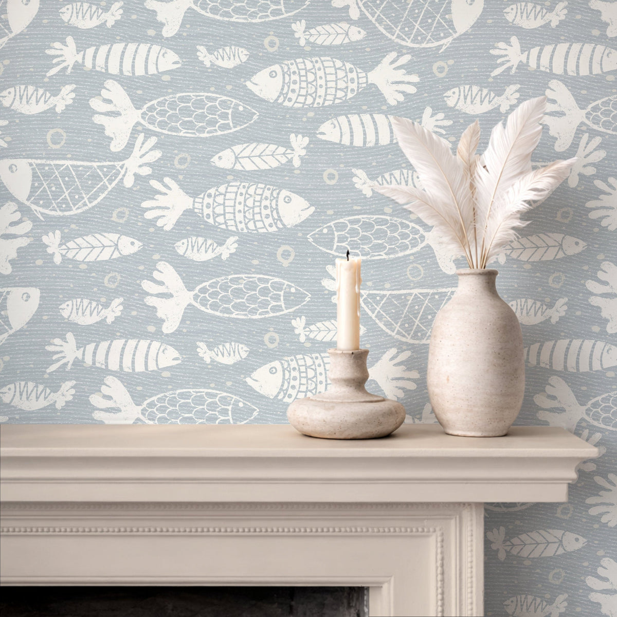 A styled interior scene showing a fireplace mantel with patterned blue and white school of fish wallpaper, a white feather vase, and a candle holder with a lit candle.