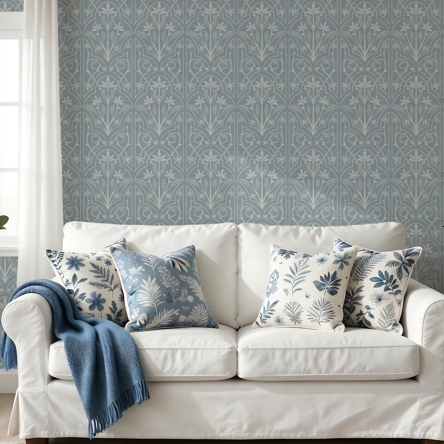 A styled living room scene featuring a white slipcovered sofa with blue and white patterned throw pillows and a blue throw blanket, set against a wall with matching blue and white art nouveau trellis wallpaper. A small wooden side table with a potted plant stands to the right.