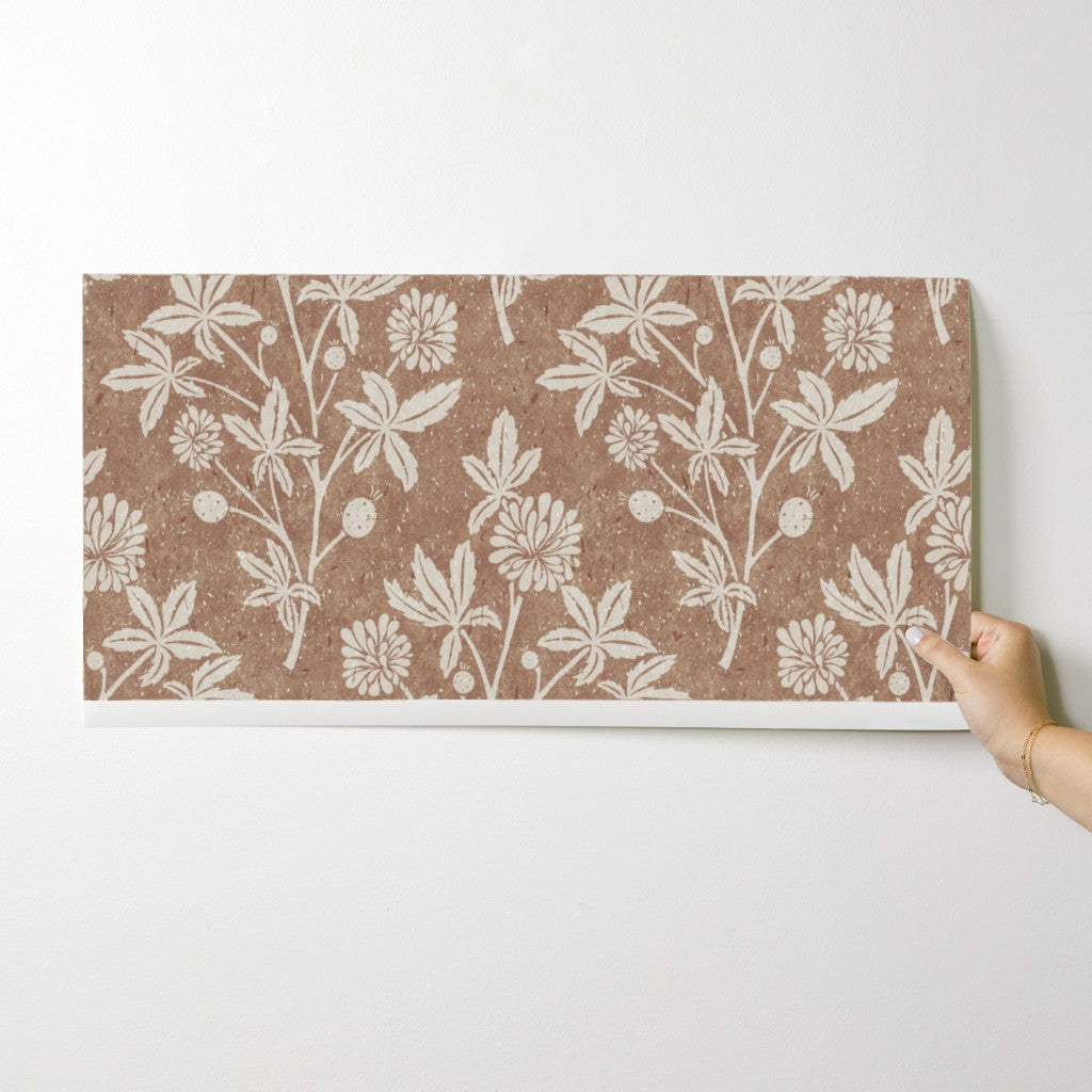 Hand holding a wallpaper swatch showing tan block print wallpaper with floral and geometric diamond pattern.