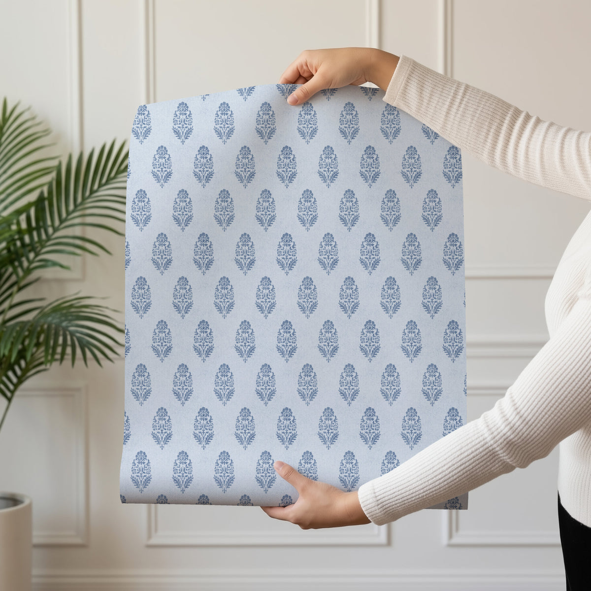 A person in a cream ribbed jumper holds up a sheet of blue floral block print wallpaper in front of a white panelled wall. A green palm plant stands on the left, and the small stylised flowers repeat in offset rows across the sheet.