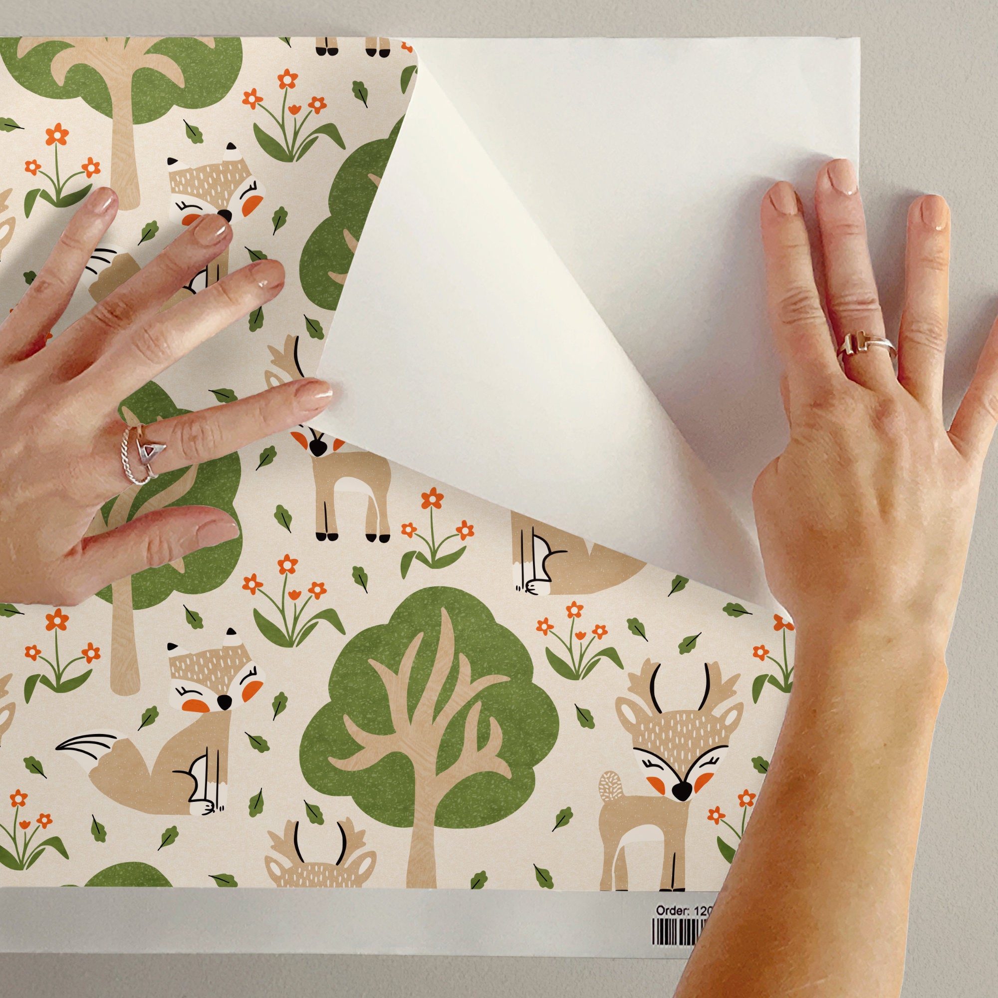 A person's hands are shown peeling back the top layer of a sample of woodland nursery wallpaper to reveal the plain white reverse side. The front layer features a whimsical forest pattern with cartoon foxes, deer, trees, and flowers on a warm cream background.