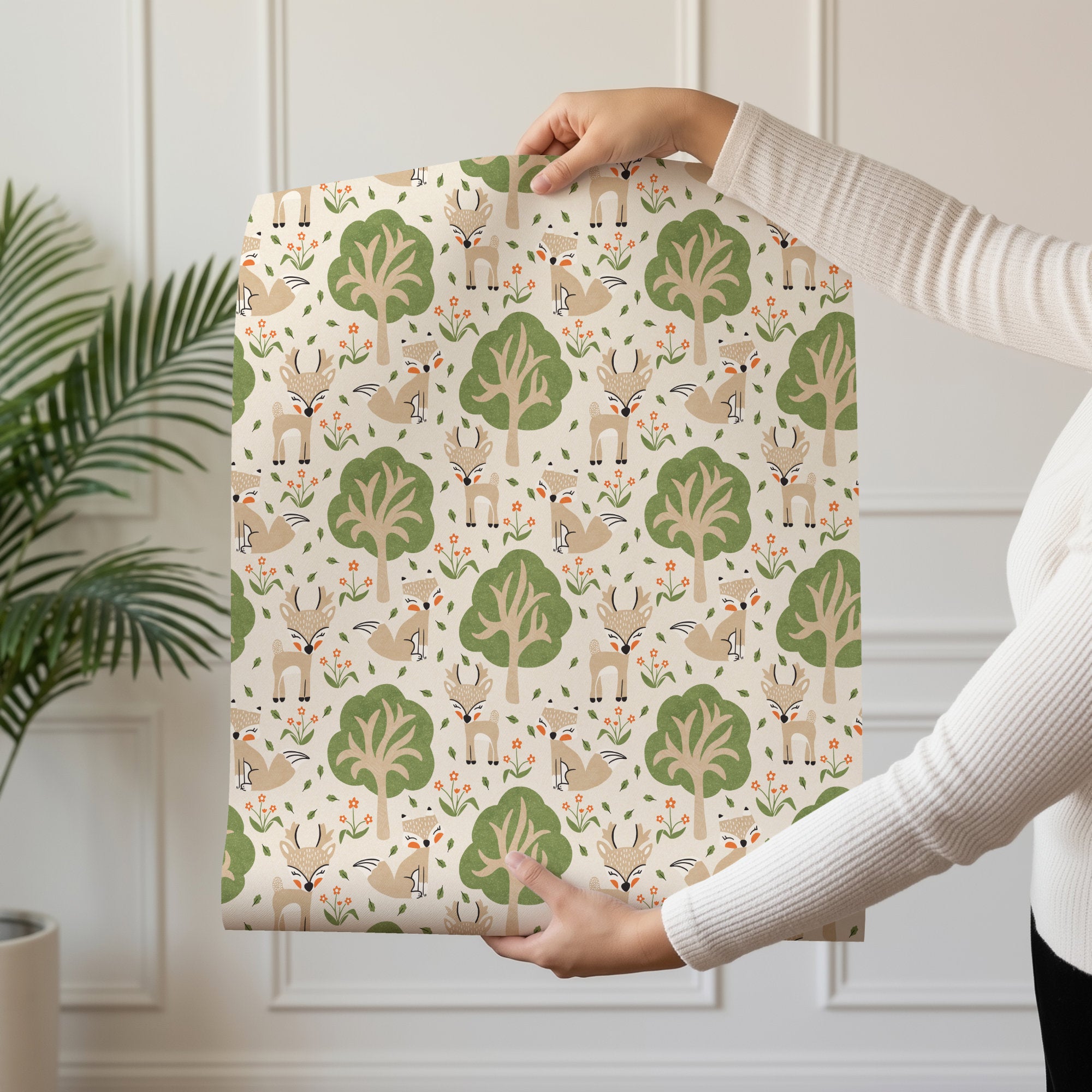 A person holds up a sheet of woodland nursery wallpaper featuring a whimsical woodland pattern of deer, foxes, trees, and flowers on a cream background. The scene is set in a bright room with a white paneled wall and a green potted plant on the left.