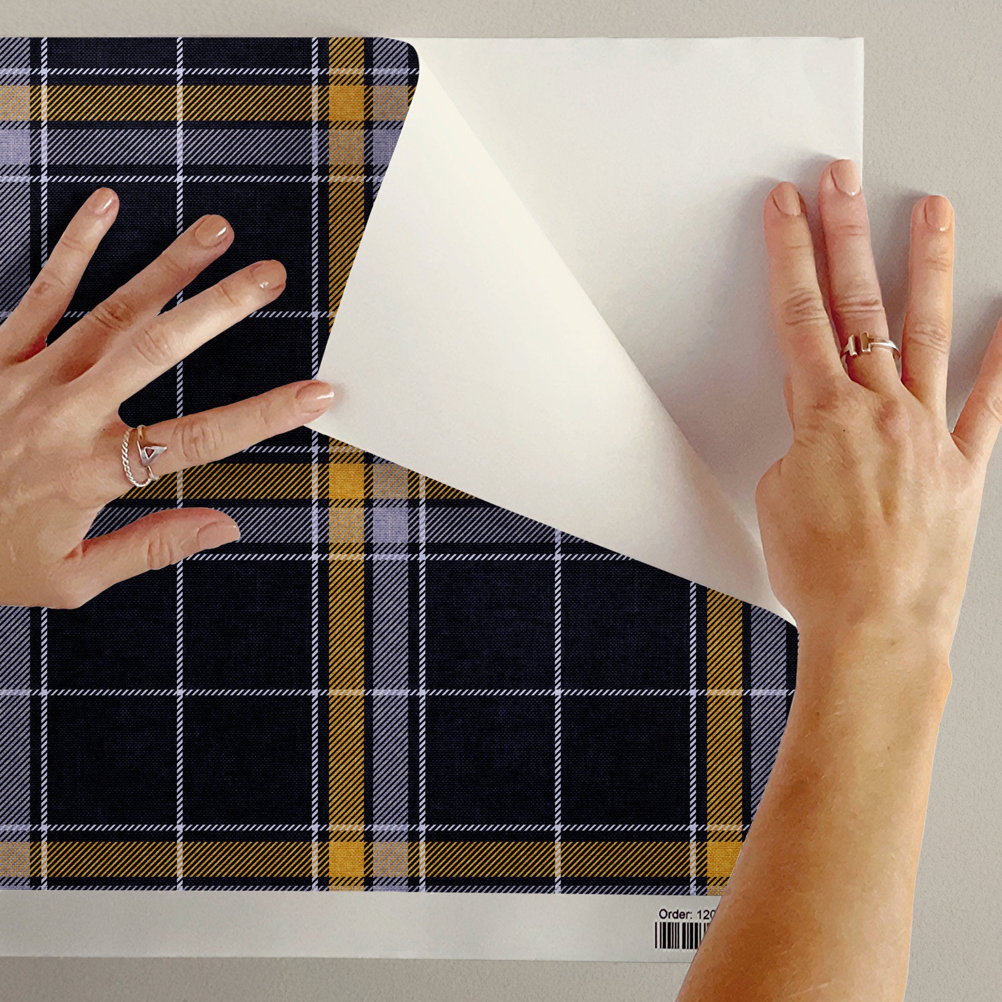A person's hands peel back a white backing from a sheet of plaid adhesive wallpaper revealing a navy blue, yellow, and white heritage plaid wallpaper pattern underneath.