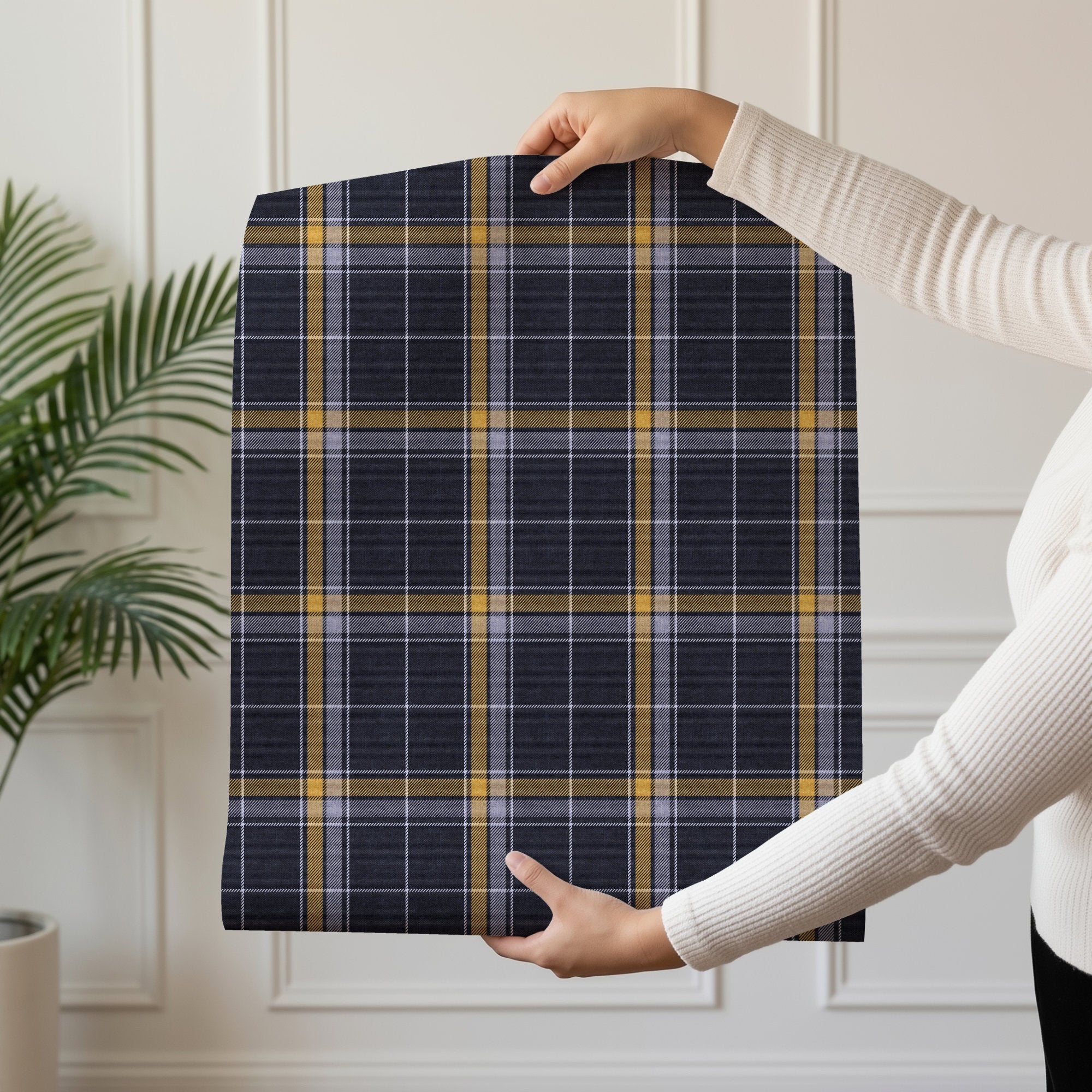 A person wearing a white ribbed long-sleeve top holds up a roll of heritage plaid wallpaper with a navy blue, gray, and mustard yellow plaid pattern in an indoor setting with a white paneled wall and a green plant.