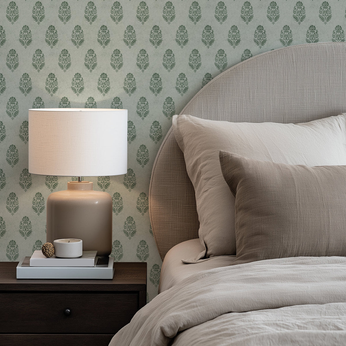 A bedroom corner with olive green floral block print wallpaper behind a bed with beige linen bedding and a rounded upholstered headboard. A white-shaded table lamp stands on a dark wood nightstand on the left, next to a stack of books.