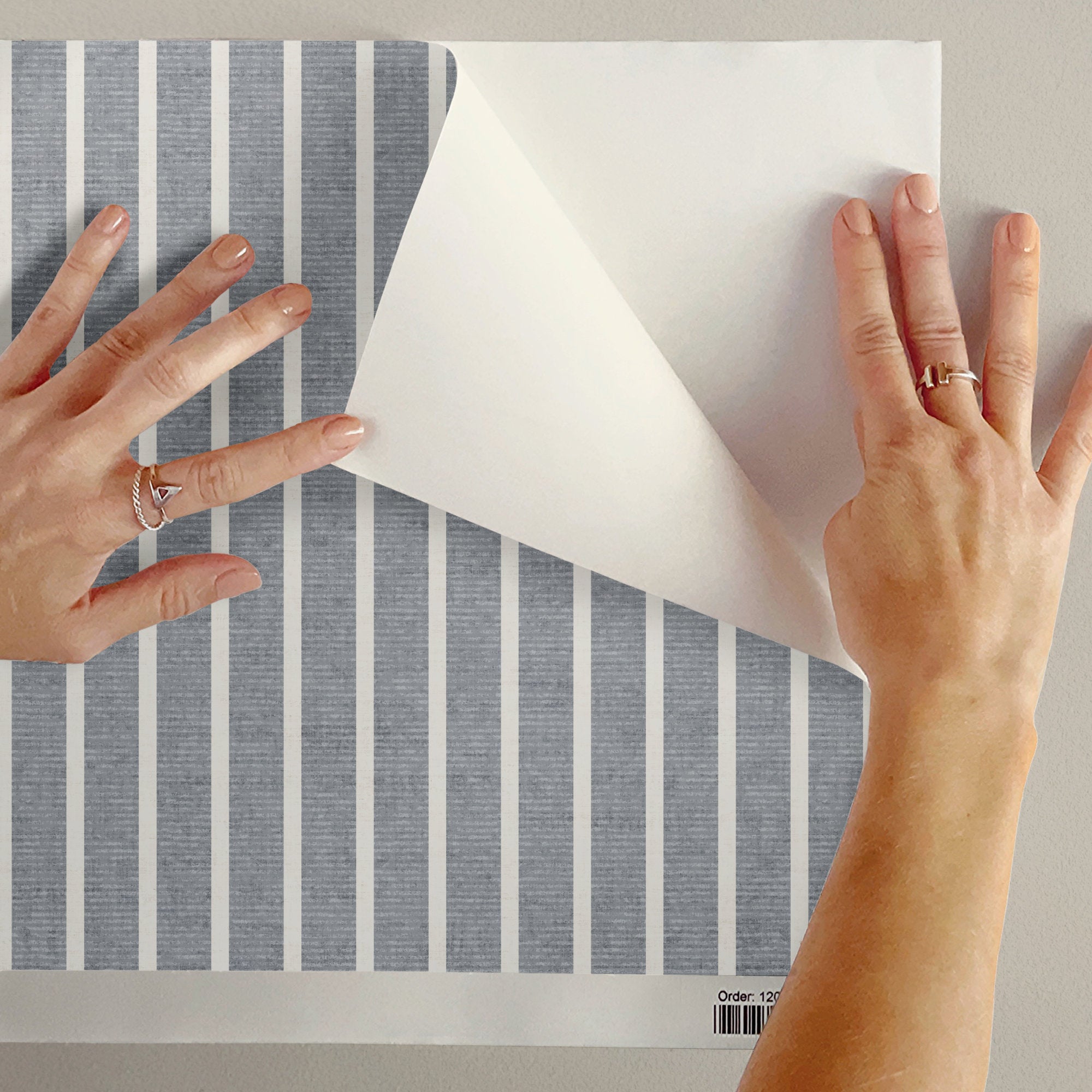 Hands peeling back the backing of light grey peel and stick pinstripe wallpaper