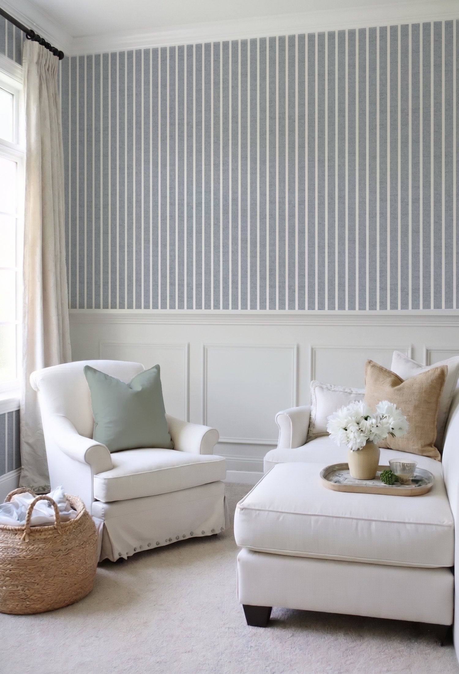 A styled living room corner featuring a white slipcovered armchair with a sage green pillow, a matching white chaise ottoman with beige pillows and a floral centerpiece, and a woven basket on a light carpet. The wall behind is decorated with gray and white vertical pinstripe wallpaper above white wainscoting.