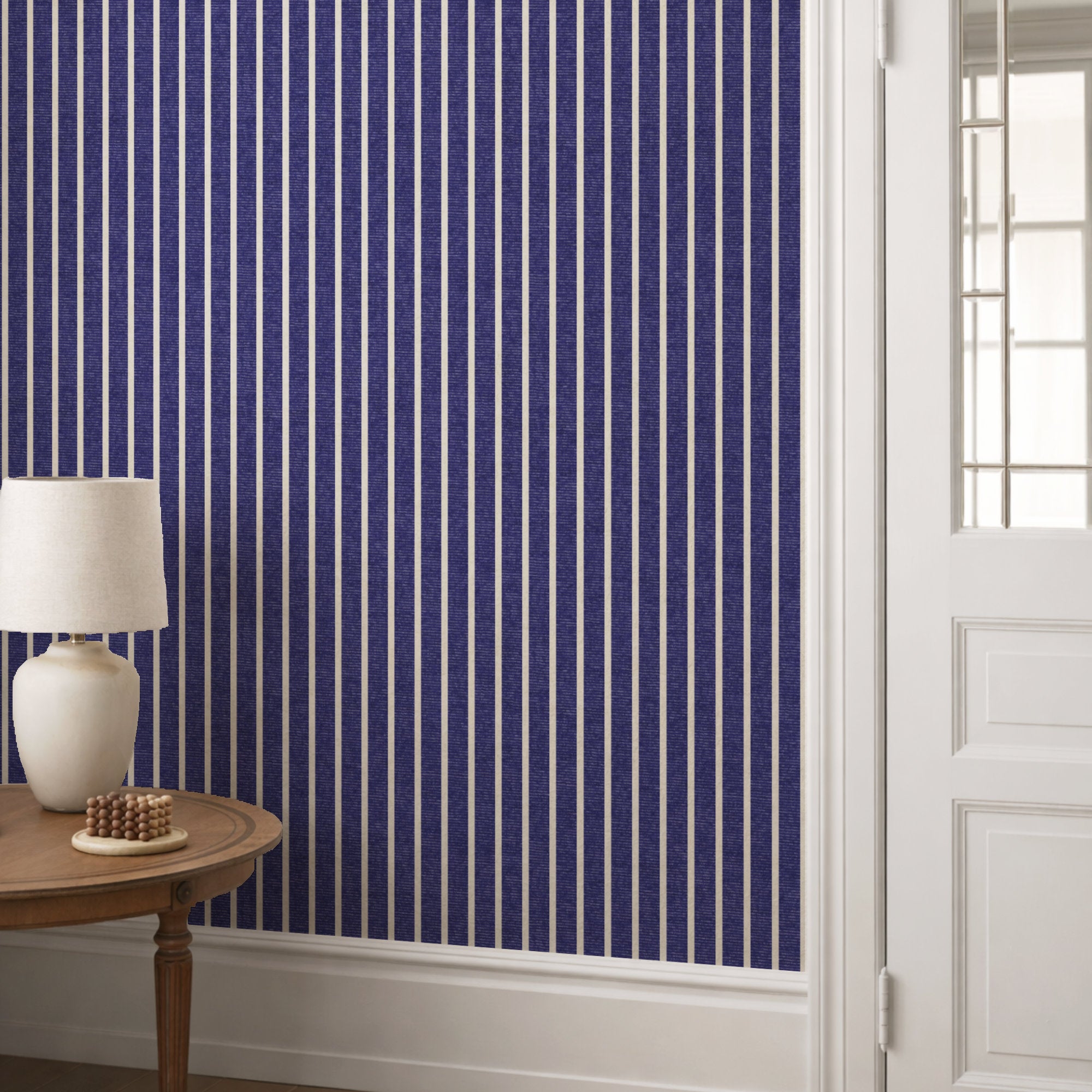 Indigo blue pinstripe wallpaper in a hallway behind a round console table with a cream lamp