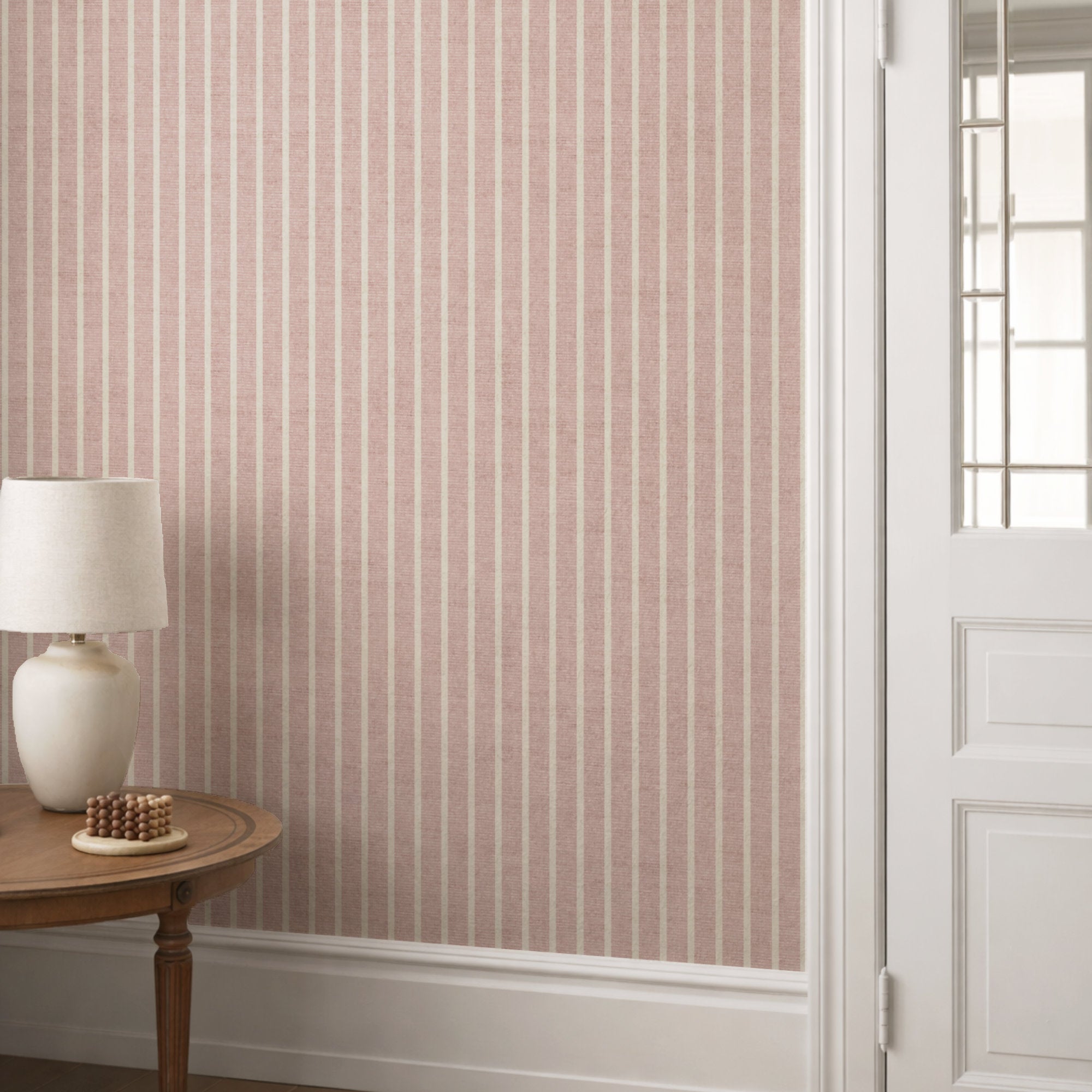 Dusty pink pinstripe wallpaper in a hallway behind a round console table with a cream lamp