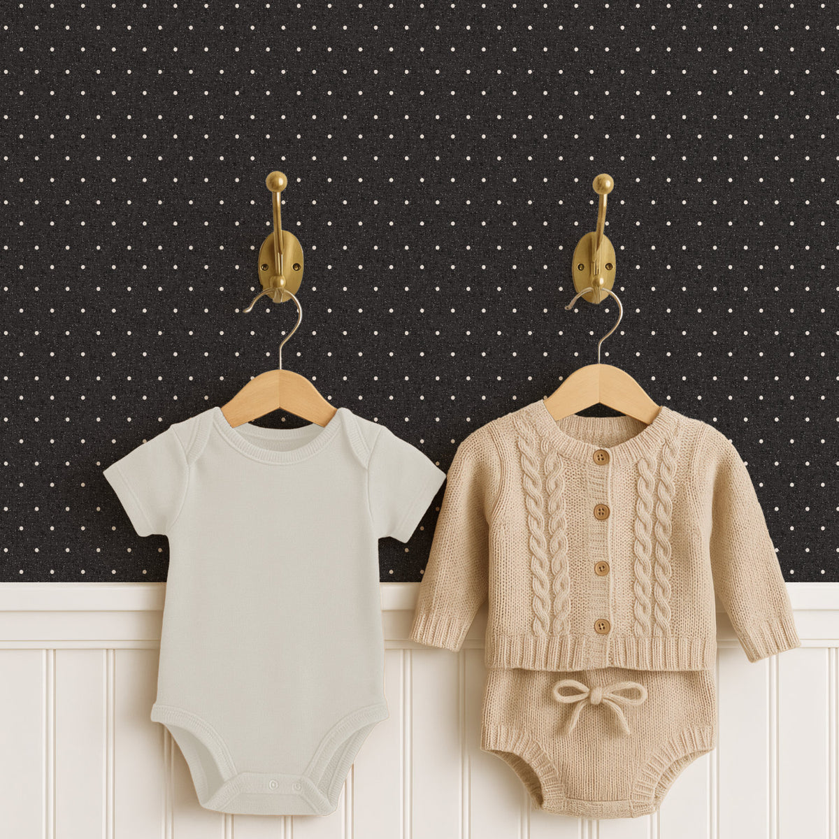 Two brass hooks hold a pale cream baby bodysuit and an oatmeal cable-knit cardigan with matching knitted shorts on wooden hangers, above white panelled wainscoting. The wall behind is near-black polka dot wallpaper, the small cream dots standing out sharply.