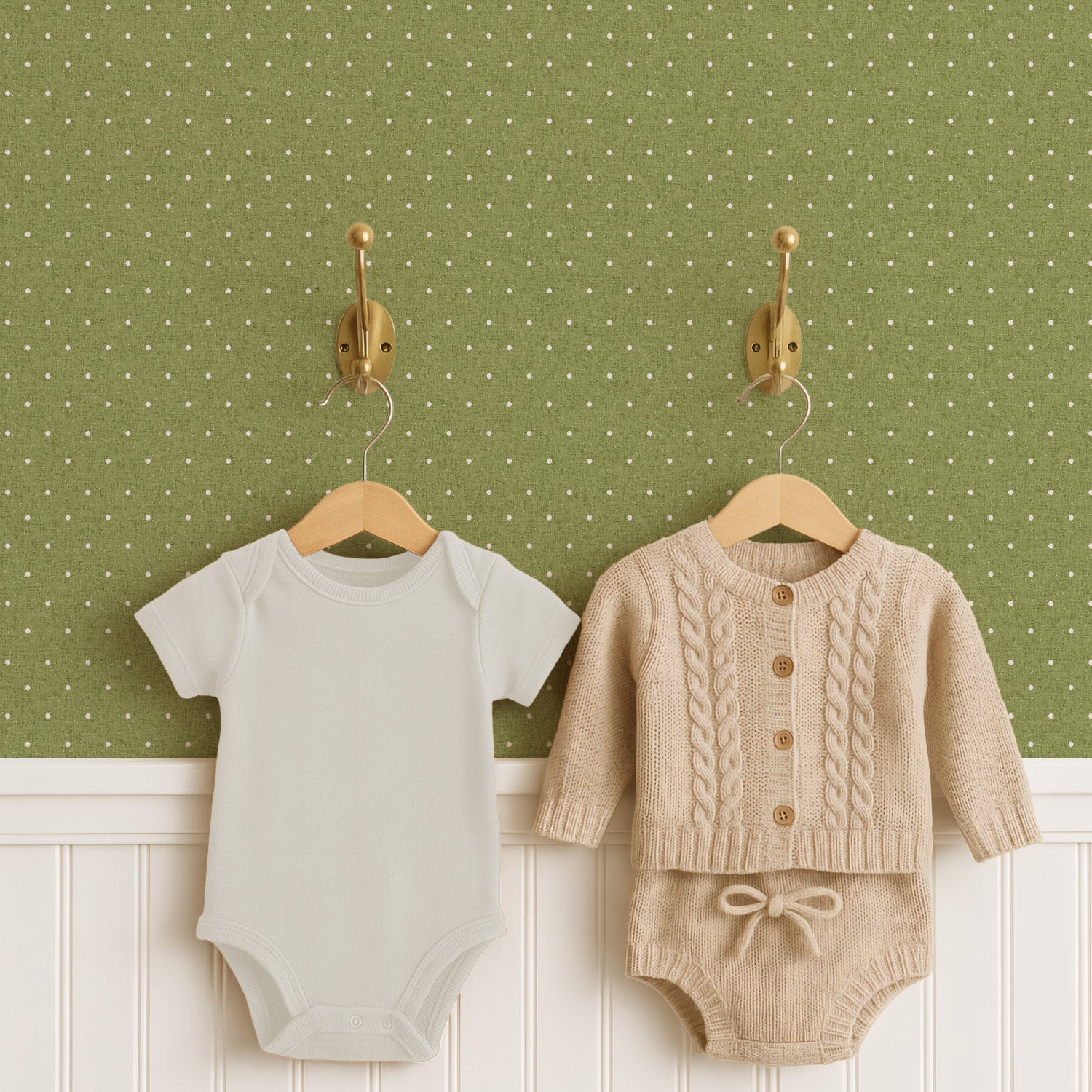 Two brass hooks hold a pale cream baby bodysuit and an oatmeal cable-knit cardigan with matching knitted shorts on wooden hangers, above white panelled wainscoting. The wall behind is olive green polka dot wallpaper scattered with small cream dots.