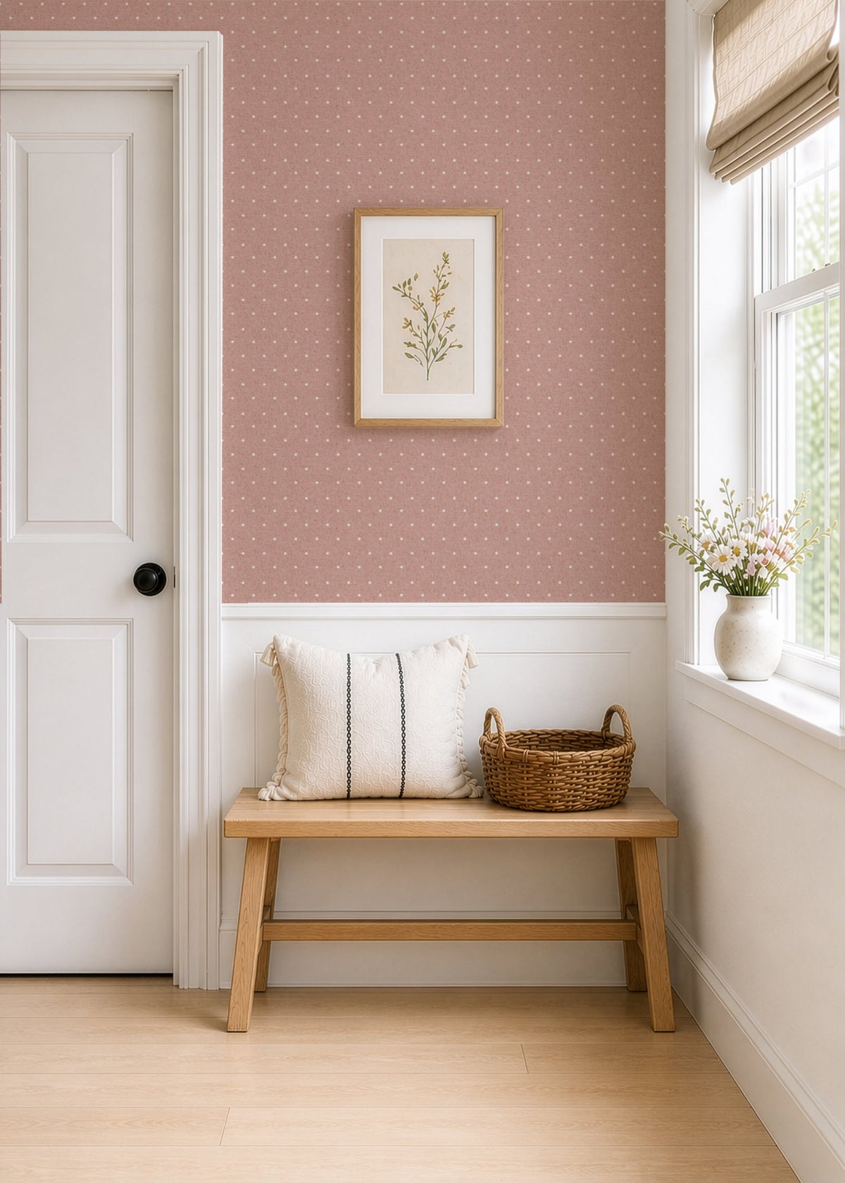 A bright hallway papered in dusty pink polka dot wallpaper, with a white panelled door on the left and a window on the right letting in daylight. A small framed botanical print hangs above a light wood bench, on a pale wood floor.