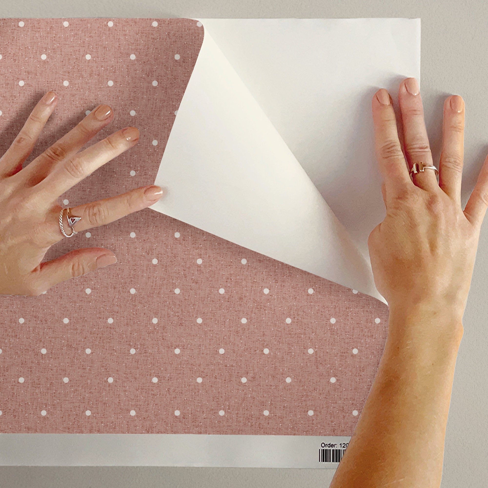 Two hands peel back the corner of a length of dusty pink polka dot wallpaper, revealing the white backing paper underneath. The small cream dots and the soft linen texture of the surface are visible up close.