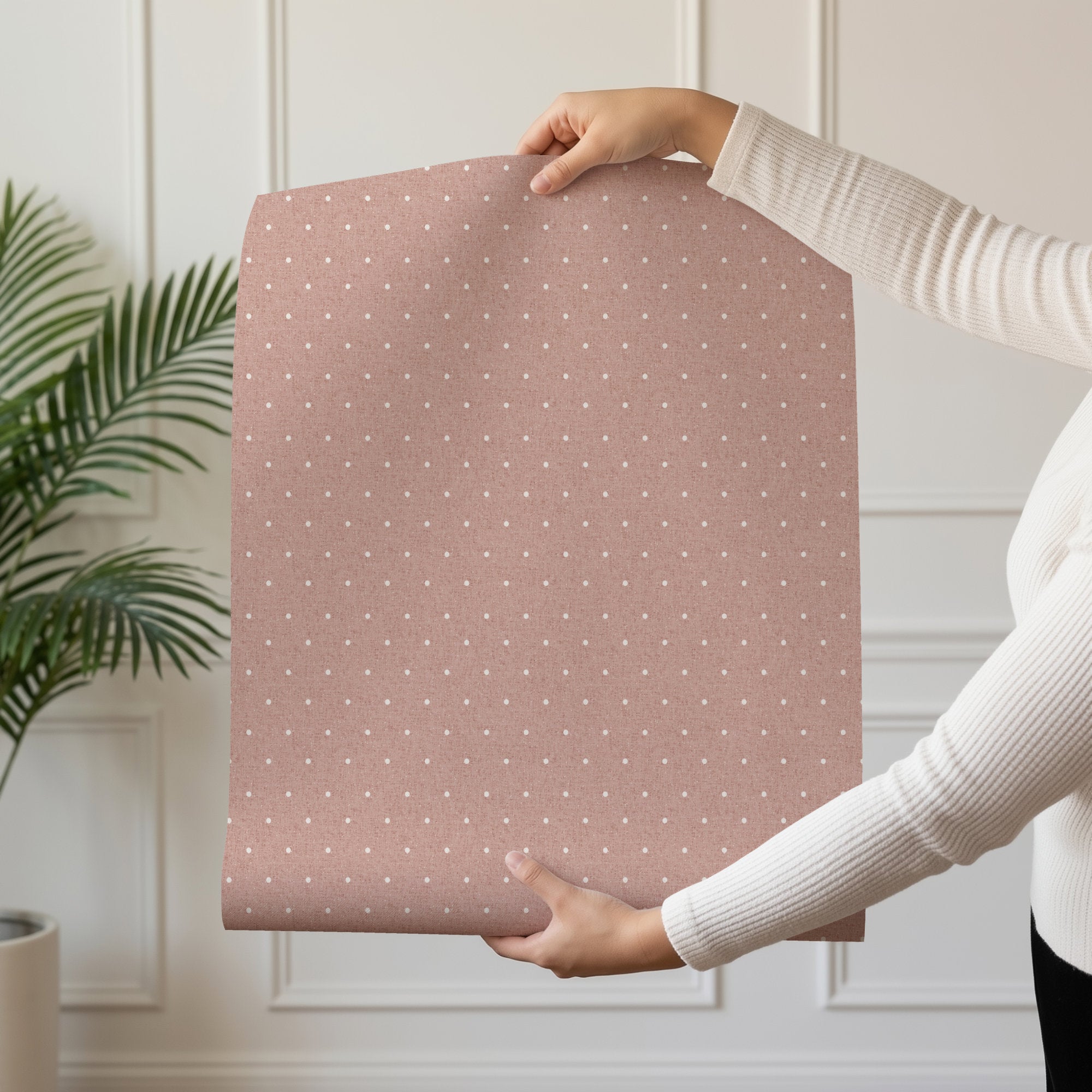 A person in a cream jumper holds up a curved sheet of dusty pink polka dot wallpaper patterned with small cream dots. Behind them is a bright room with a white panelled wall and green plant fronds at the left edge.
