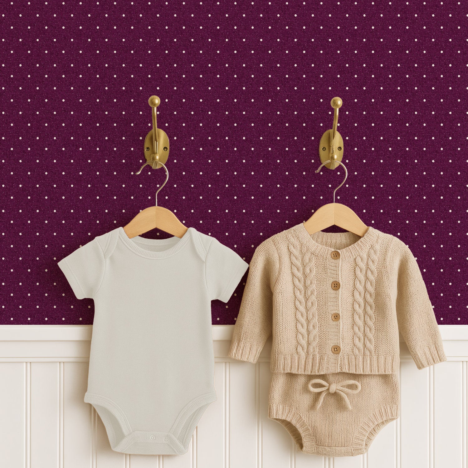 A pale cream baby bodysuit and an oatmeal cable-knit cardigan with matching knitted shorts hang from two brass hooks on wooden hangers, above white panelled wainscoting. The wall behind is deep plum polka dot wallpaper, the small cream dots bright against the dark ground.