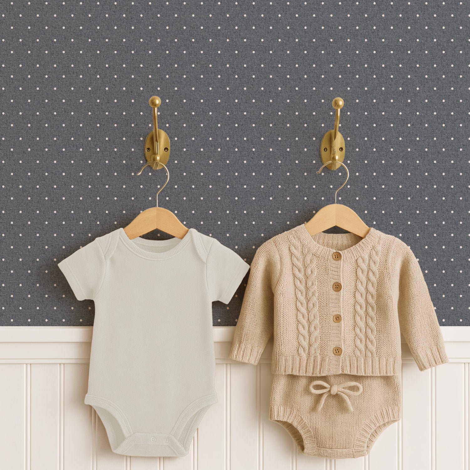 Two brass hooks hold a pale cream baby bodysuit and an oatmeal cable-knit cardigan with matching knitted shorts on wooden hangers, above white panelled wainscoting. Behind them, slate grey polka dot wallpaper carries small cream dots in an even grid.