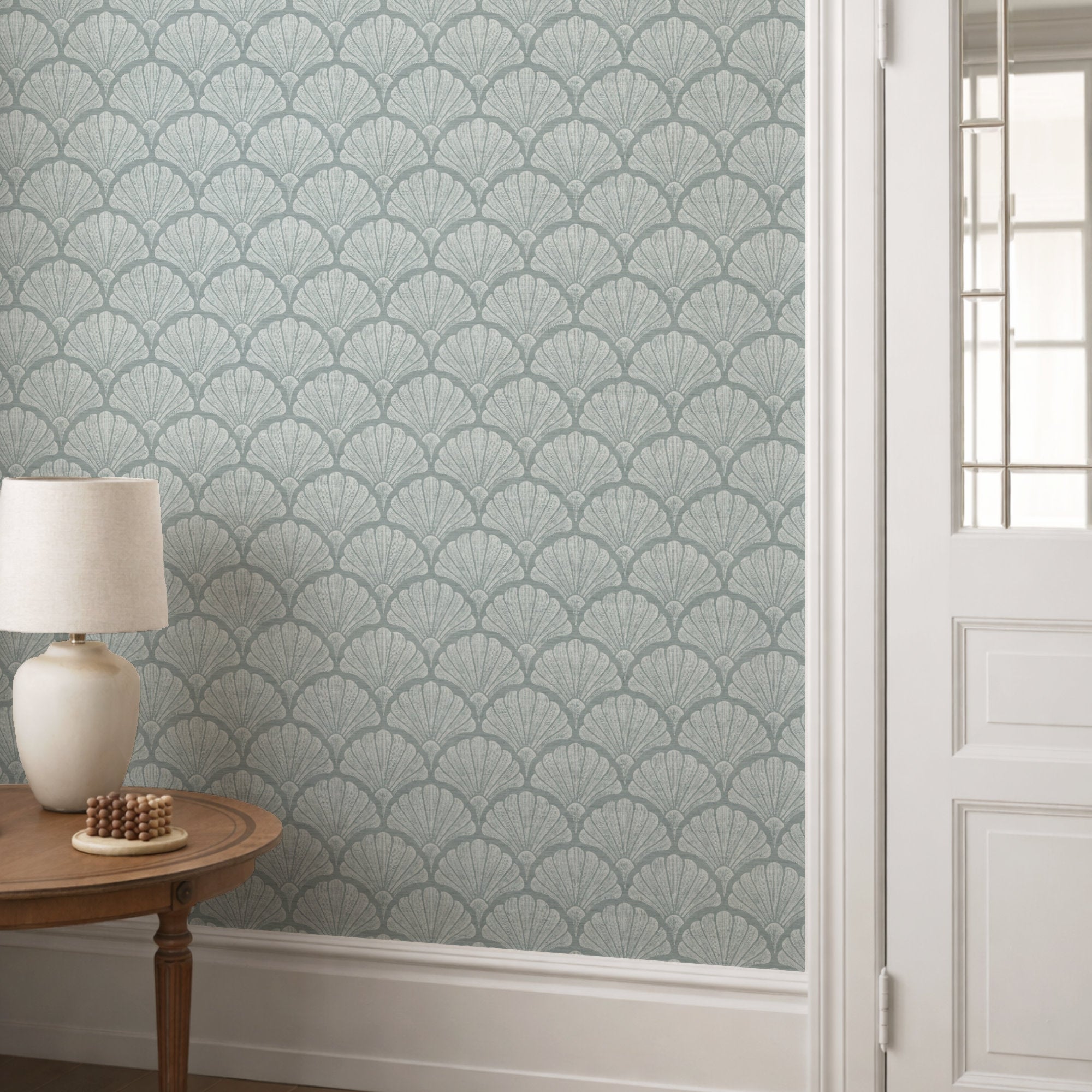 Light blue scallop shell wallpaper in a hallway, hand-drawn fan shells behind a small round table and lamp beside a panelled door