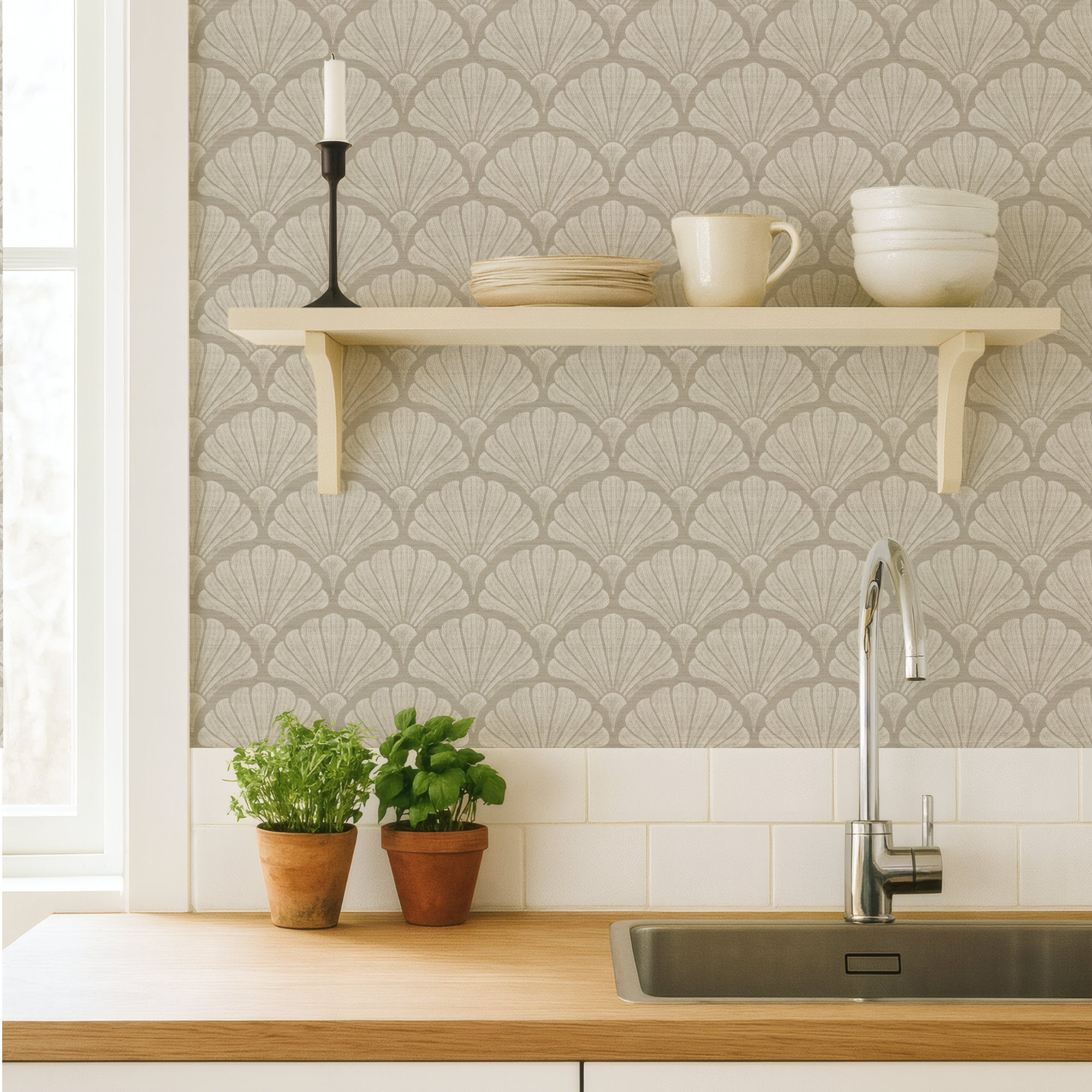 Sandy beige scallop shell wallpaper in a kitchen, hand-drawn fan shells behind a cream shelf of crockery, above a wood worktop, sink and potted herbs