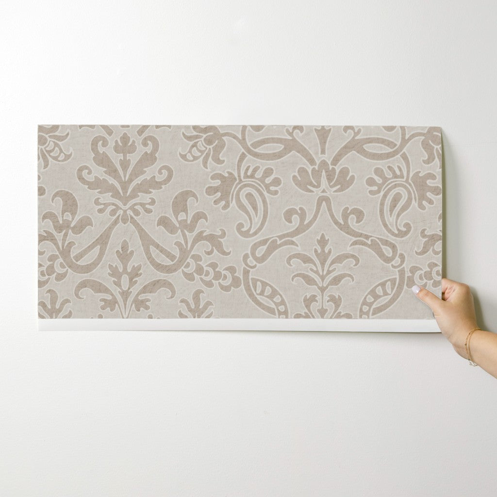 Hand holding a neutral Victorian damask floral wallpaper sample against a white wall.