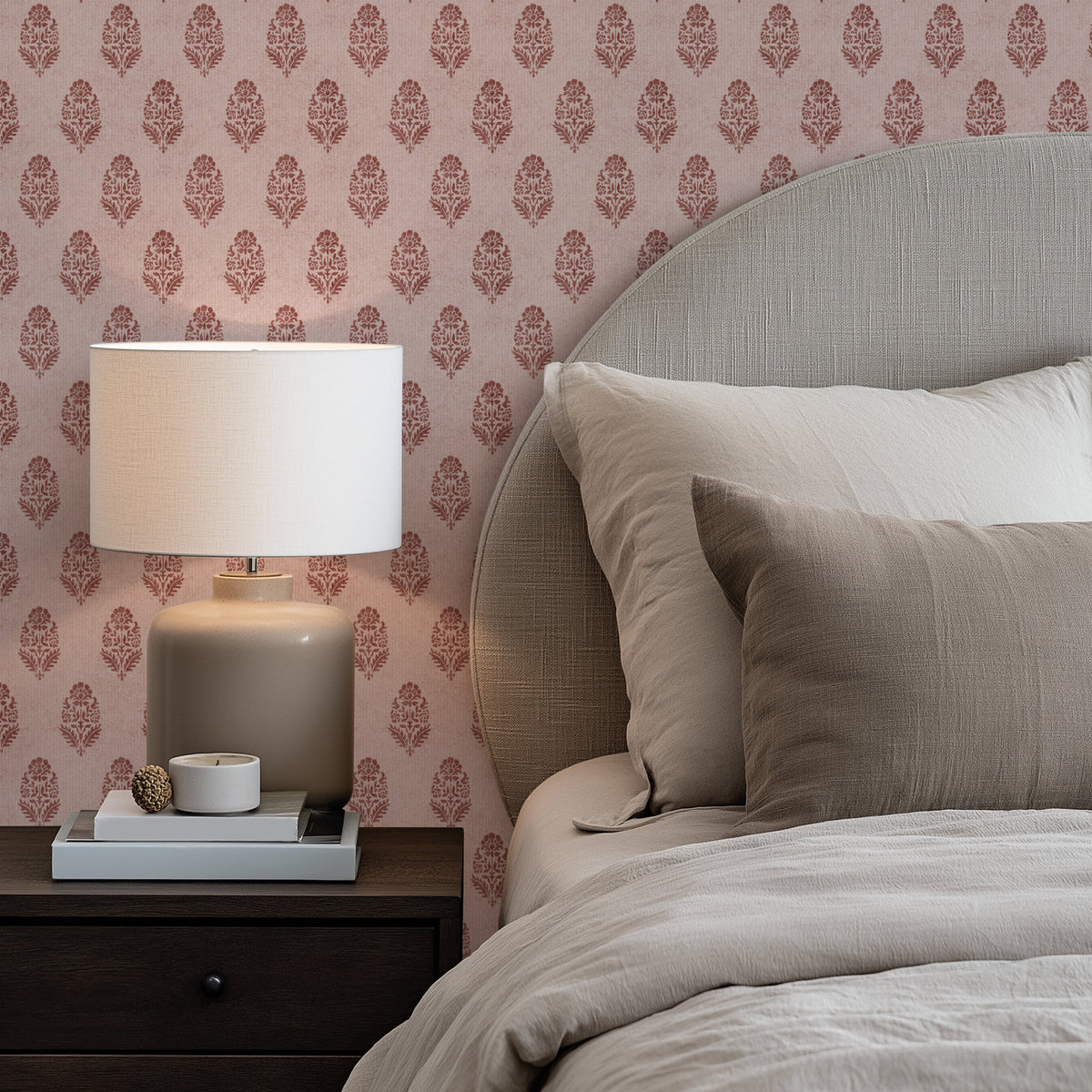 A bedroom corner with terracotta red floral block print wallpaper behind a bed dressed in soft beige bedding, with a rounded upholstered headboard. On the left, a lamp with a white shade sits on a dark wood nightstand with books and a small bowl.